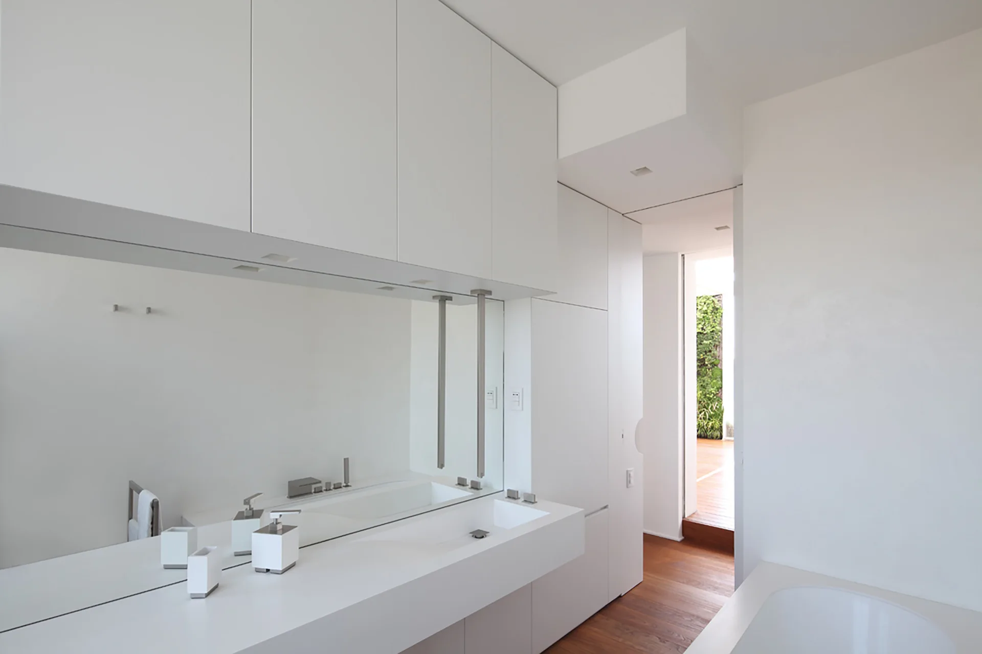A minimalist white bathroom features an integrated solid-surface sink vanity, a large wall mirror, and handleless upper cabinets.