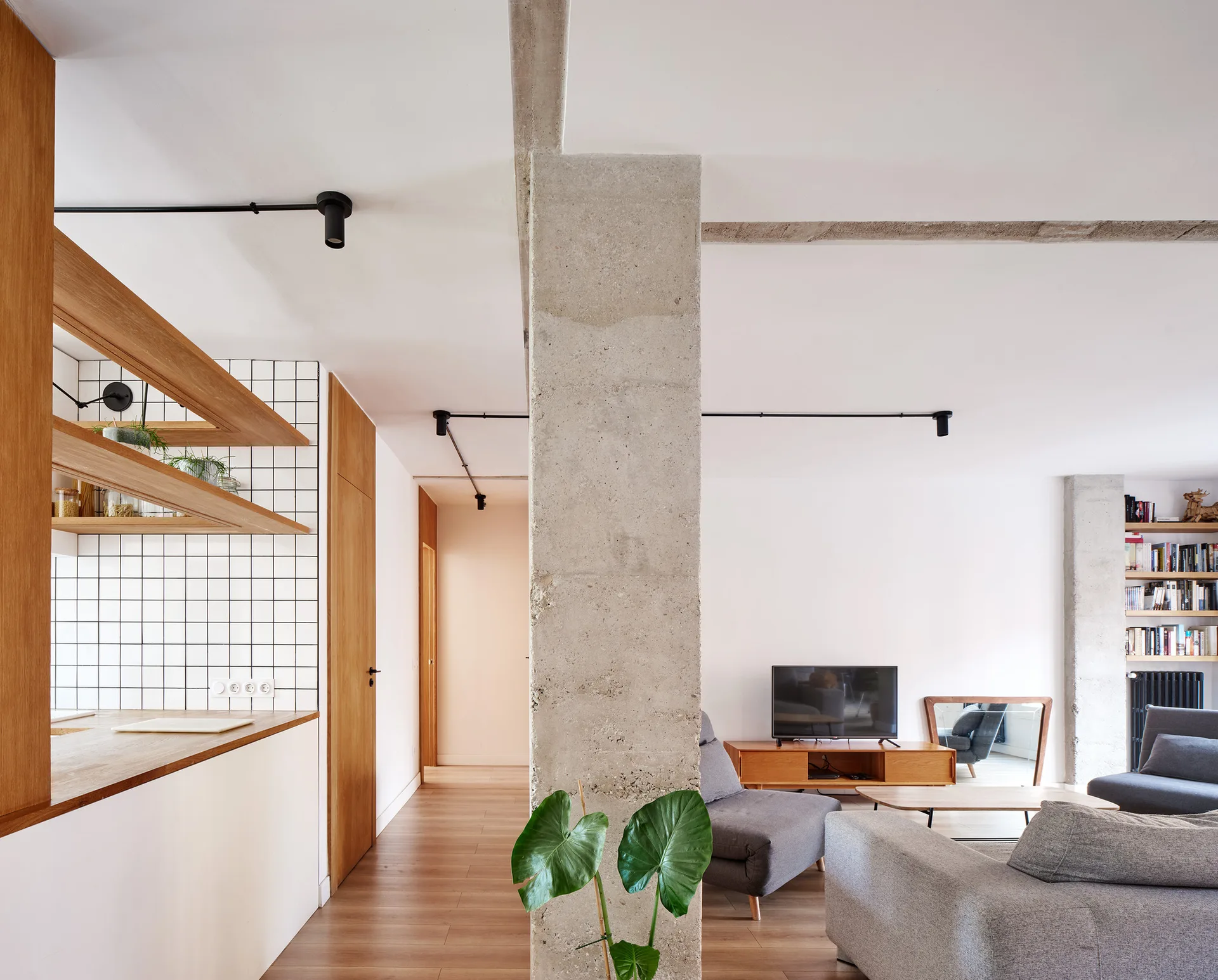 An open-plan living area showing a rough concrete column, kitchen corner with tiled walls, and a grey sofa facing a television console.