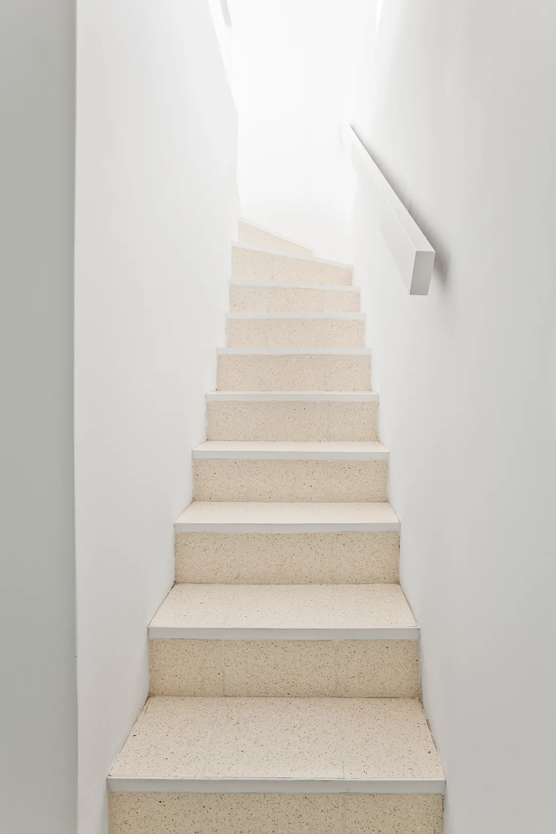 A minimalist straight staircase with light beige speckled terrazzo treads is flanked by plain white walls and a matching white handrail.