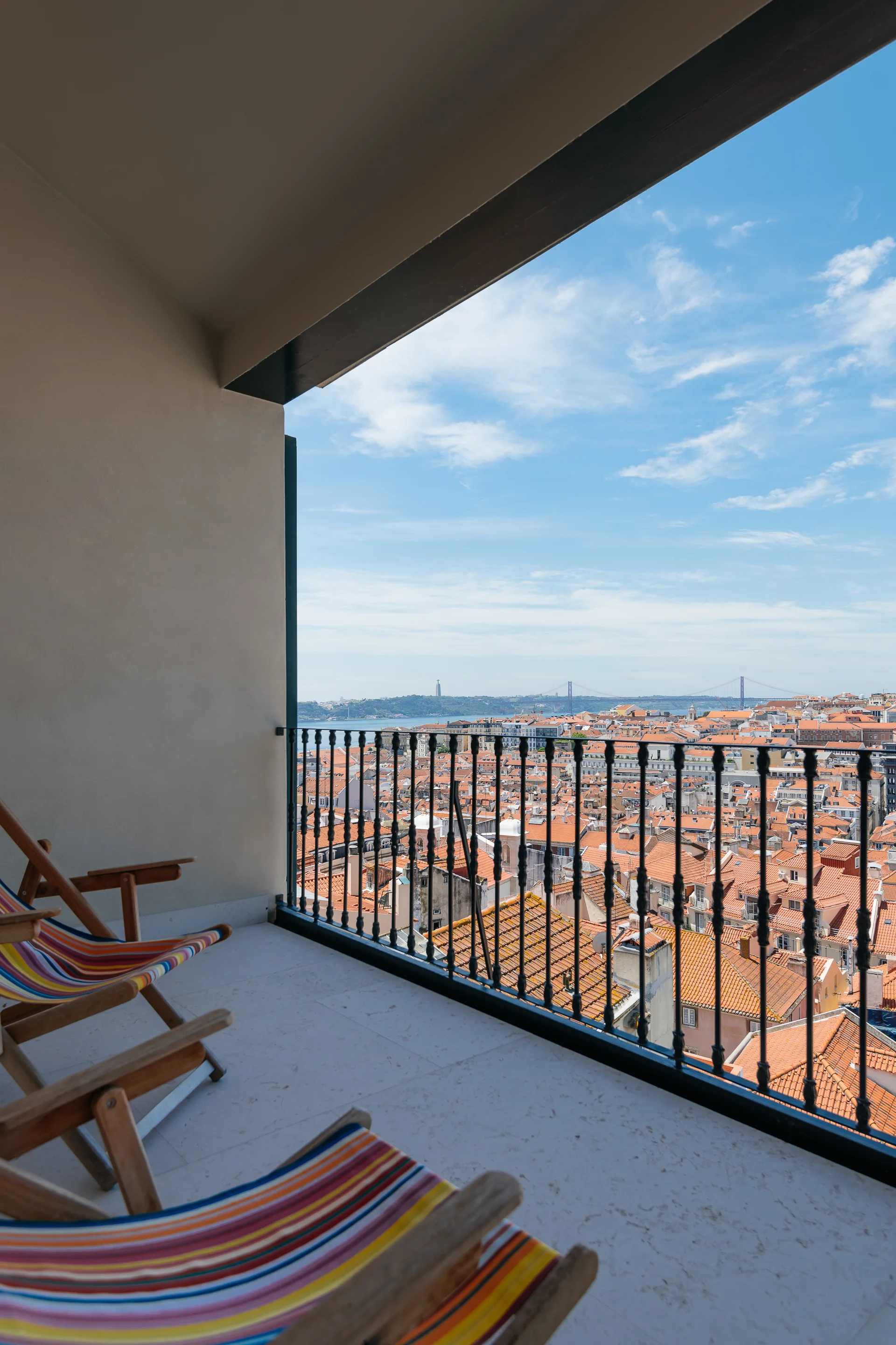 Wooden deck chairs with colorful striped fabric sit on a stone-tiled balcony overlooking a city with terracotta roofs and a bridge in the distance.