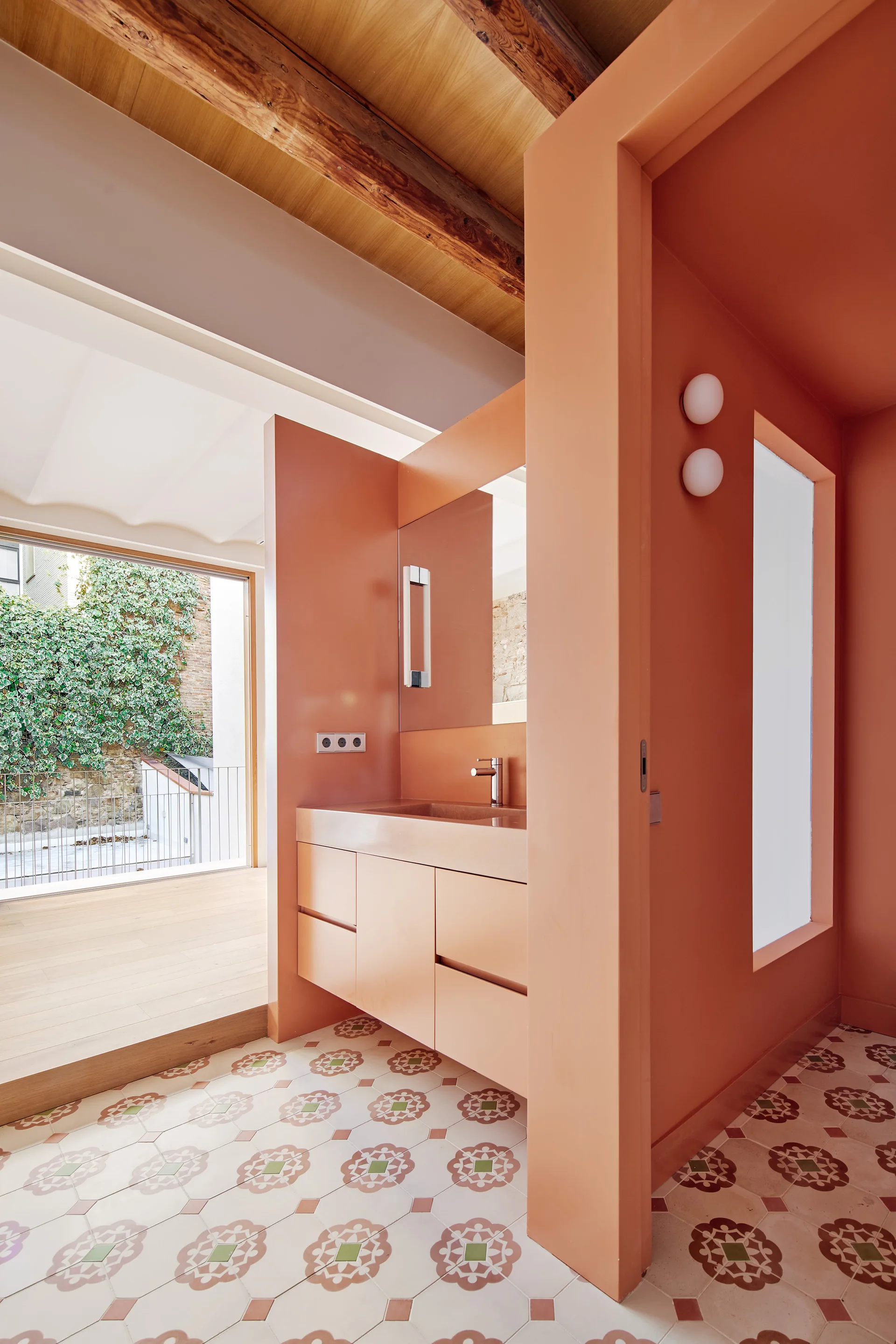 A modern terracotta bathroom features a matching floating vanity, patterned floor tiles, white round sconces, and exposed wooden ceiling beams.
