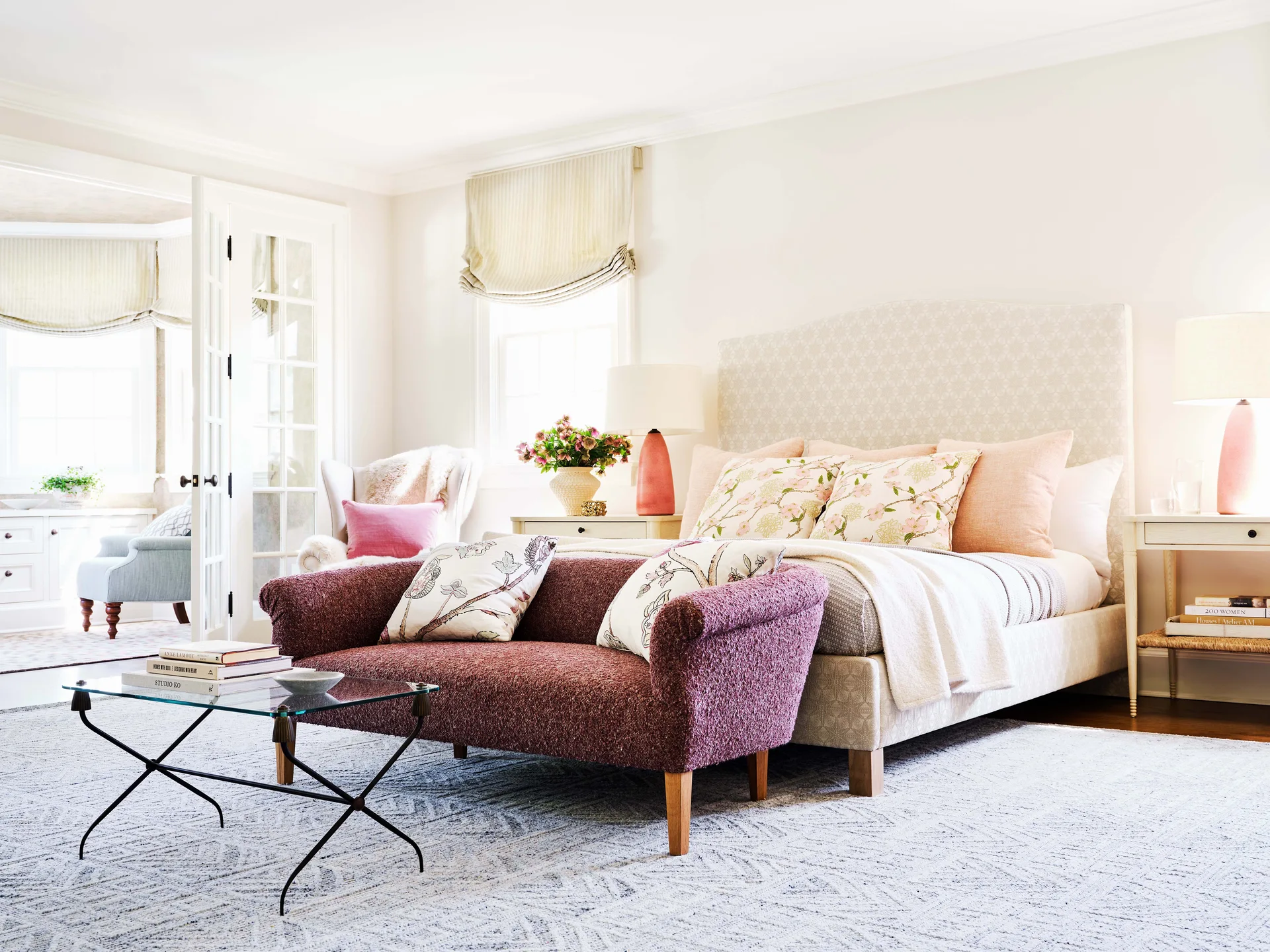 A light-filled bedroom featuring an upholstered patterned headboard, a textured purple settee at the foot of the bed, and bedside tables with pink lamps.