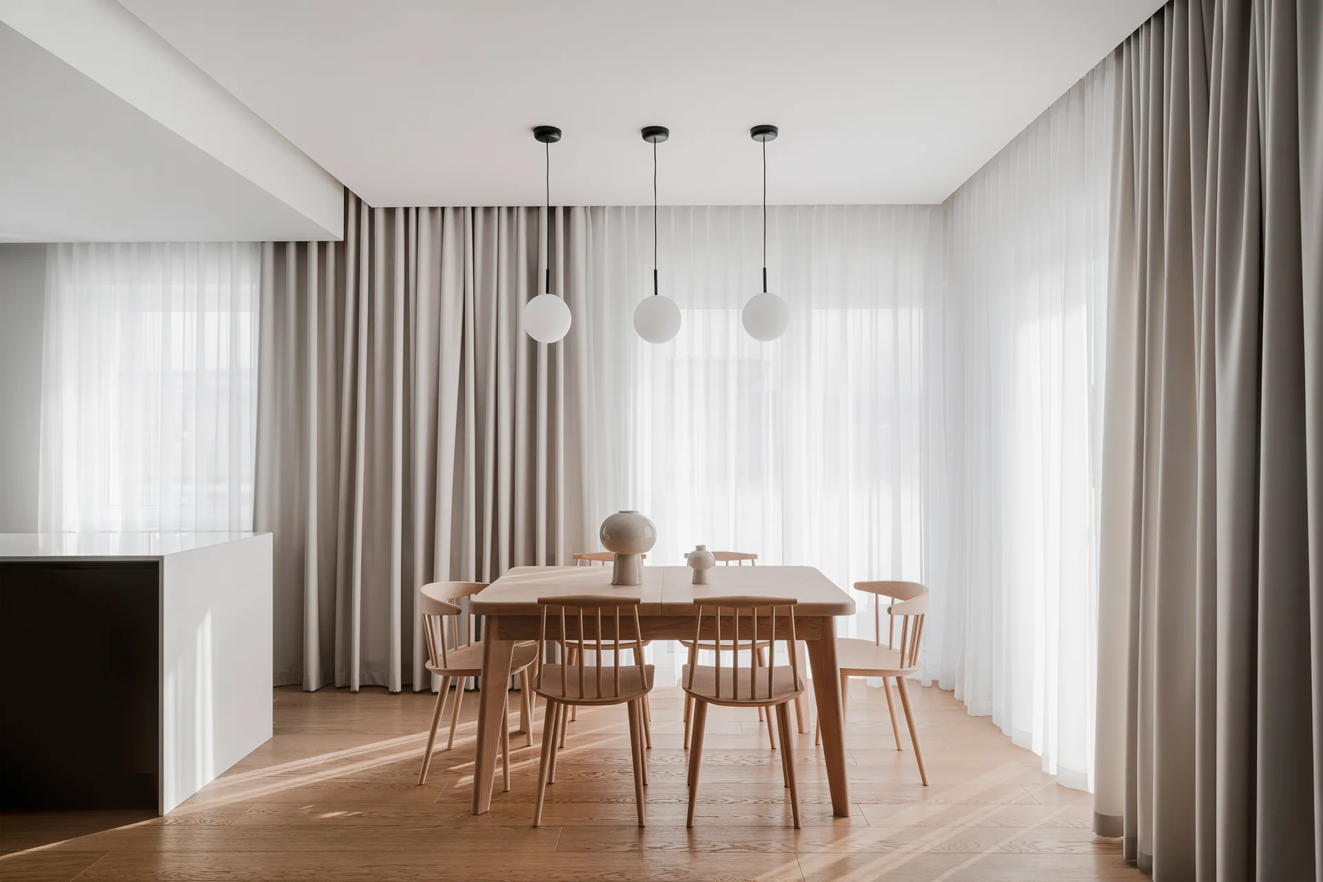 A minimalist dining room features a light wood table and spindle-back chairs beneath three globe pendant lights, set against floor-to-ceiling curtains.