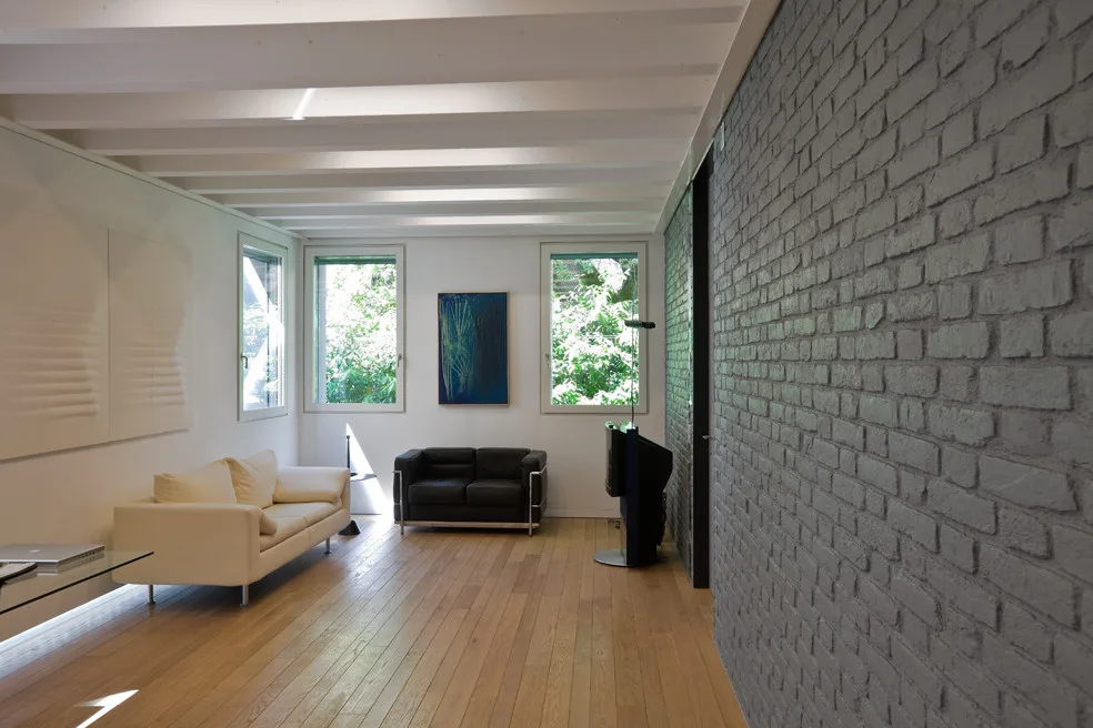 A modern living room features a textured grey painted brick wall, exposed white ceiling beams, light wood floors, and minimalist black and white sofas.