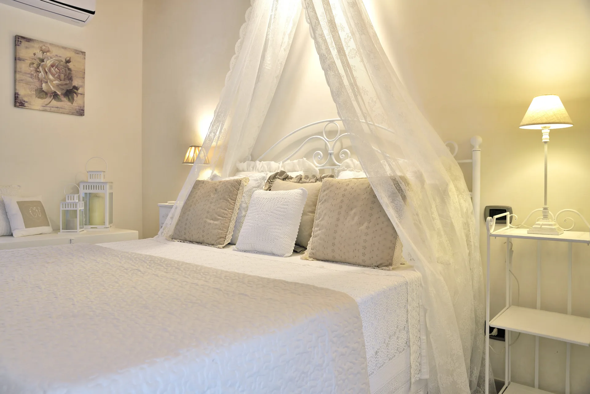A neutral bedroom features a white metal canopy bed with sheer lace drapes, decorative throw pillows, and warm bedside table lamps.