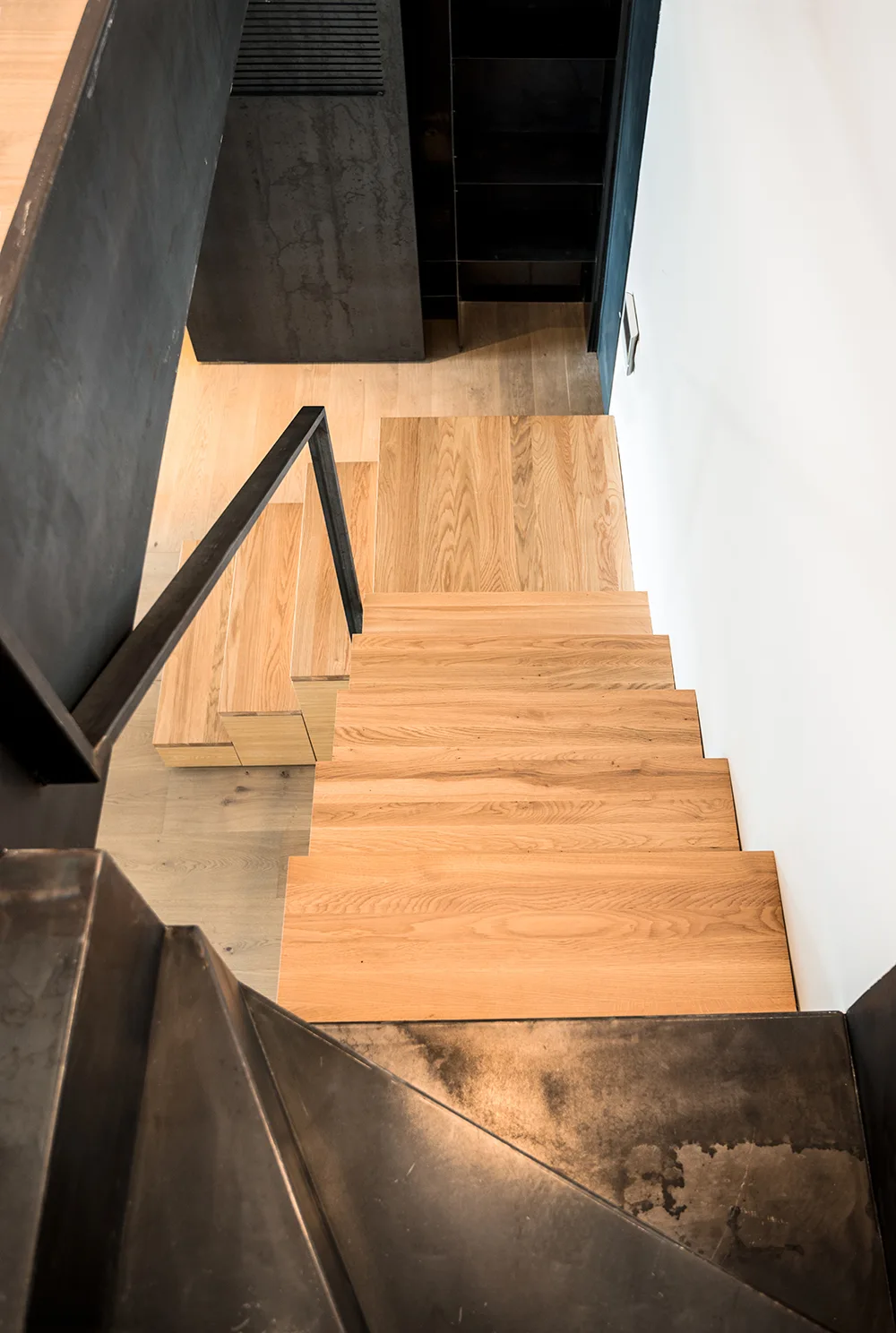 A high-angle view of a minimalist staircase featuring light oak treads, a black metal railing, and a matching dark metal landing structure.