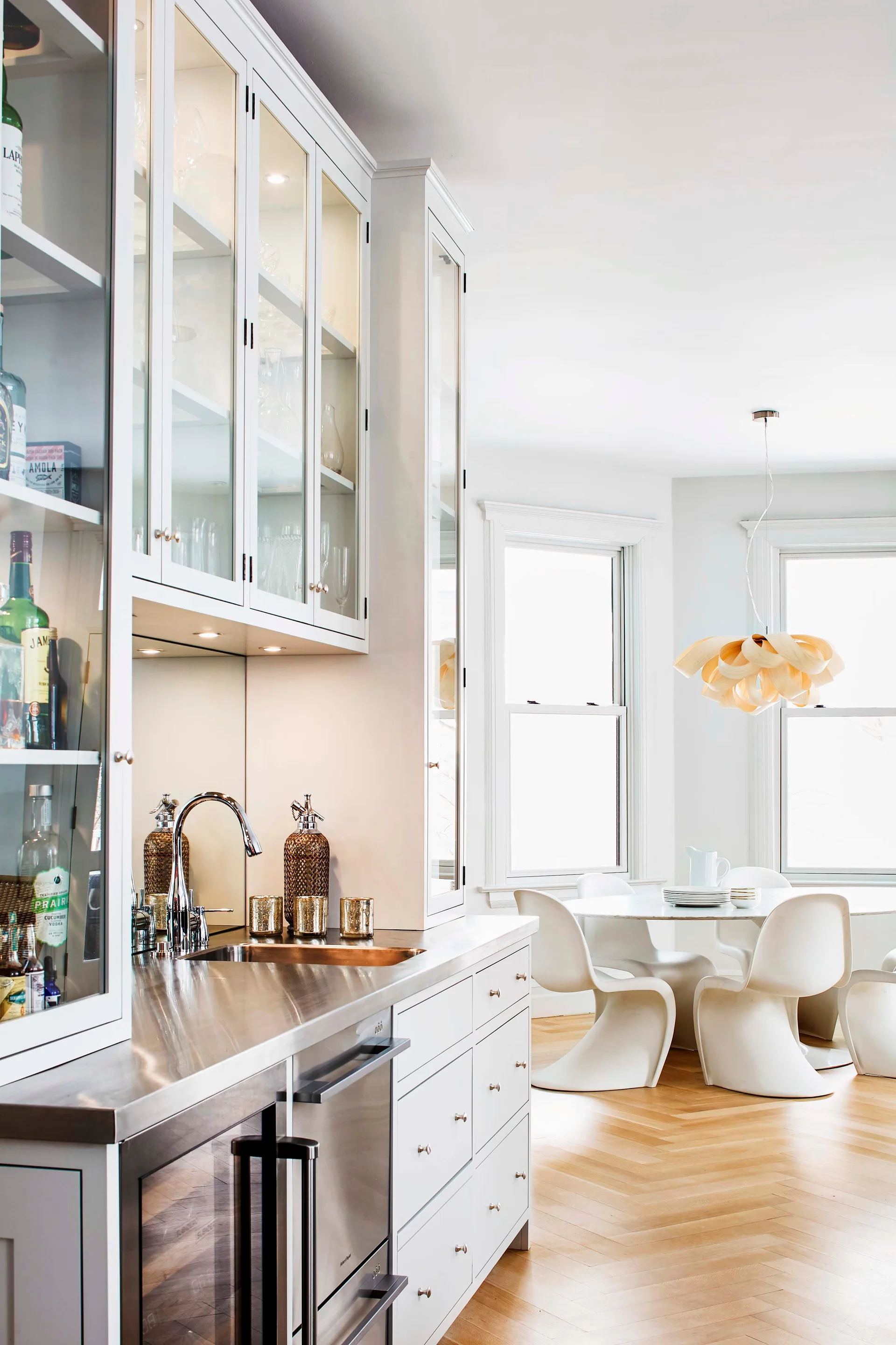 A kitchen transition zone features white cabinets with a stainless steel counter and an adjacent dining area with white sculptural chairs.