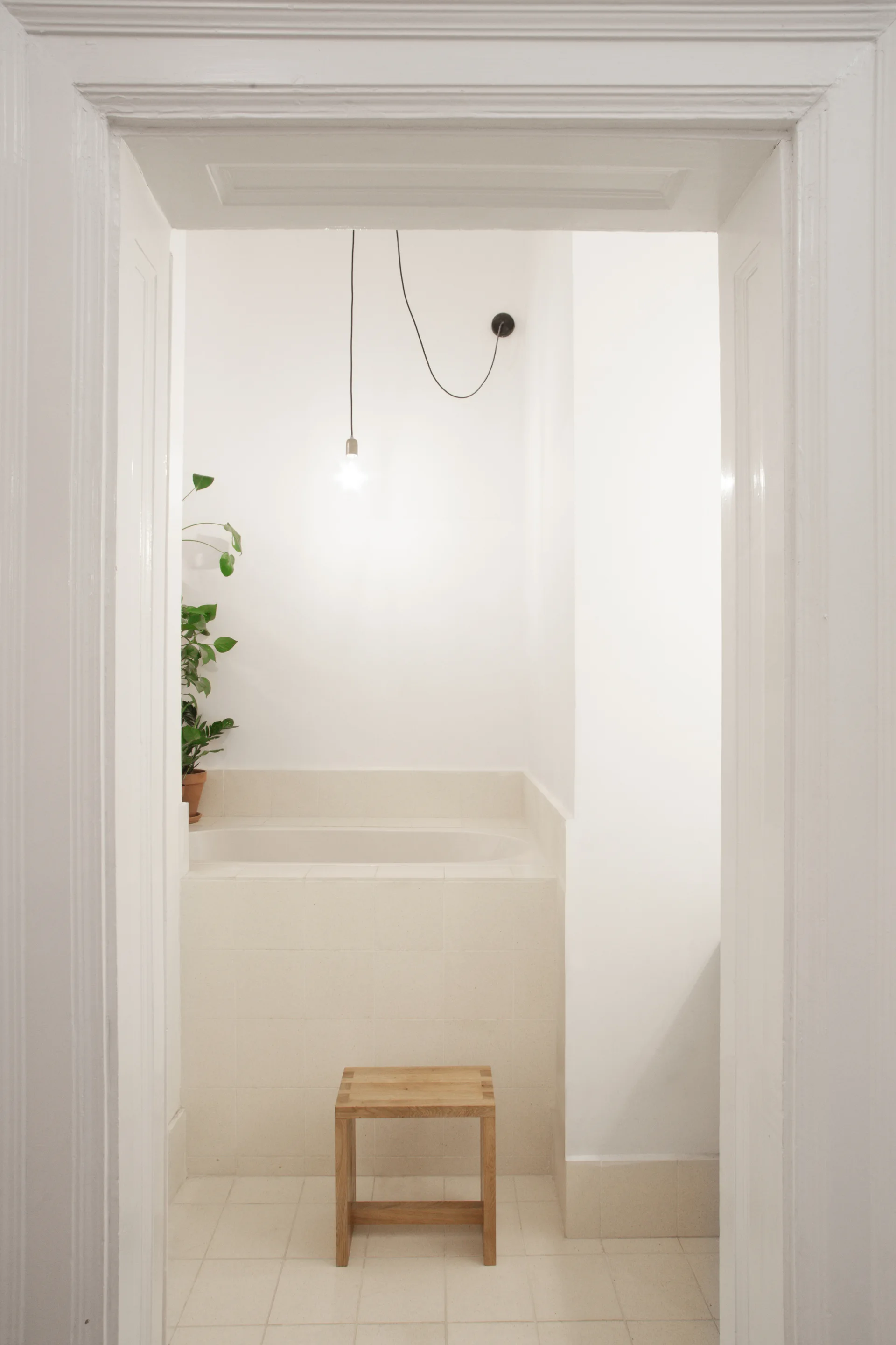 A view through a white doorway into a minimalist bathroom featuring a beige tiled bathtub, a wooden stool, and a hanging pendant bulb.