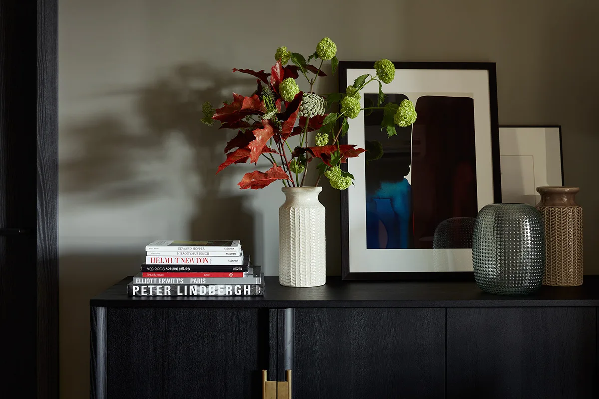 A stack of art books, a textured white ceramic vase holding leafy green branches, and framed abstract artwork sit on a dark wooden sideboard.