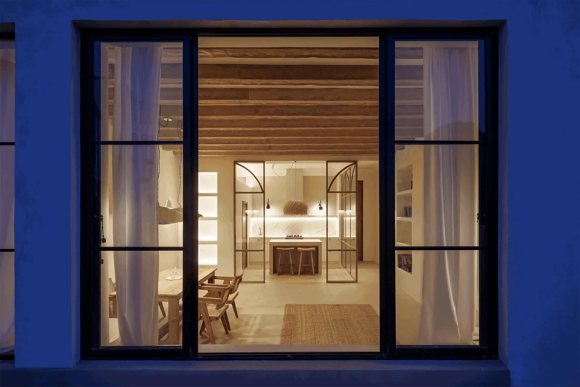 An interior view of a warm cream kitchen and dining room with exposed wood ceiling beams, seen through a dark metal window frame.