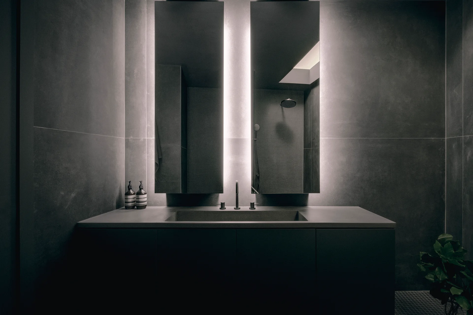 A dimly lit charcoal grey bathroom features a dark matte vanity with a three-piece faucet and two tall, narrow backlit mirrors.