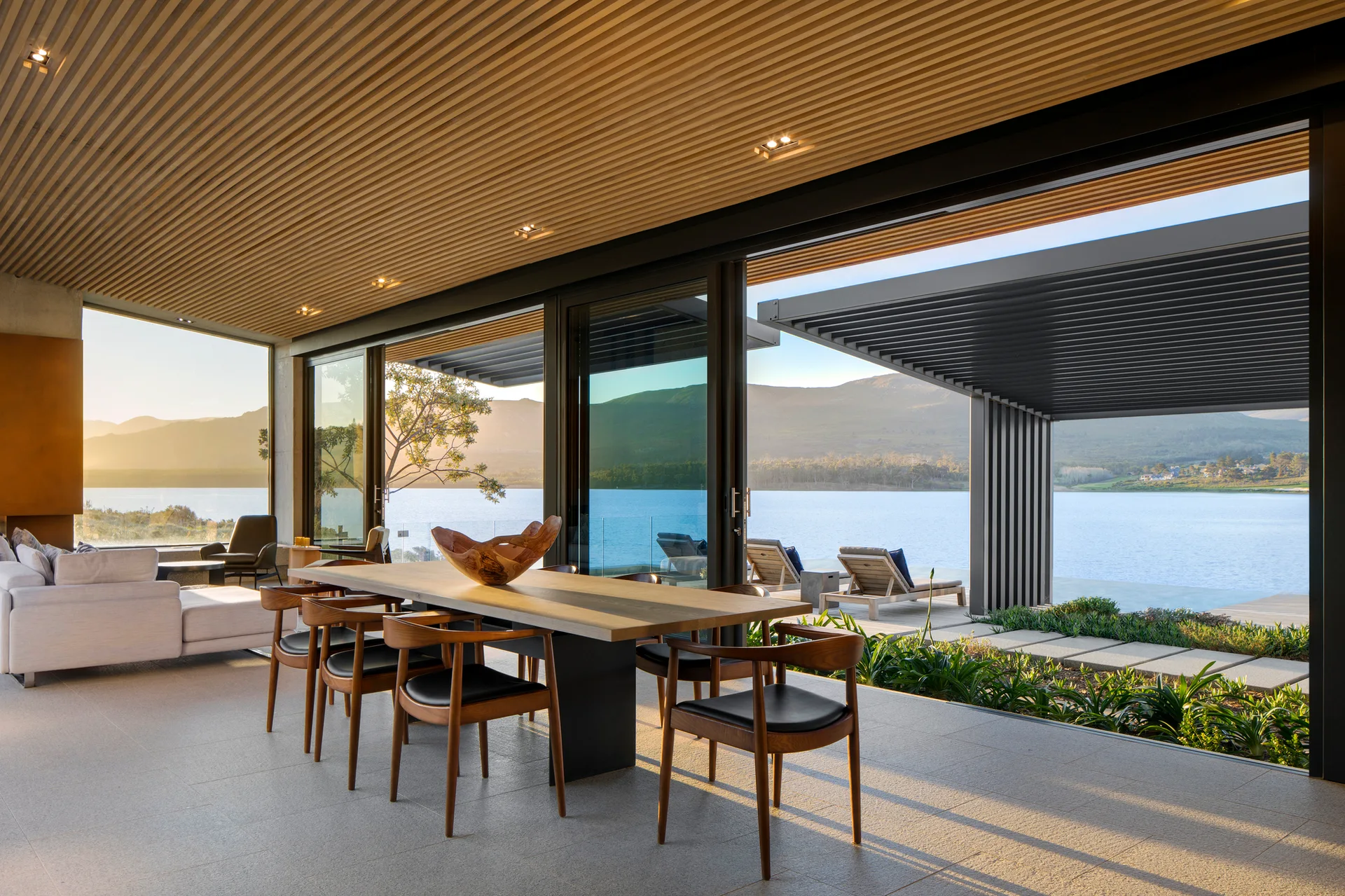 A modern dining room with a wooden table and chairs features a wood-slatted ceiling and glass sliding doors opening to a scenic lake.