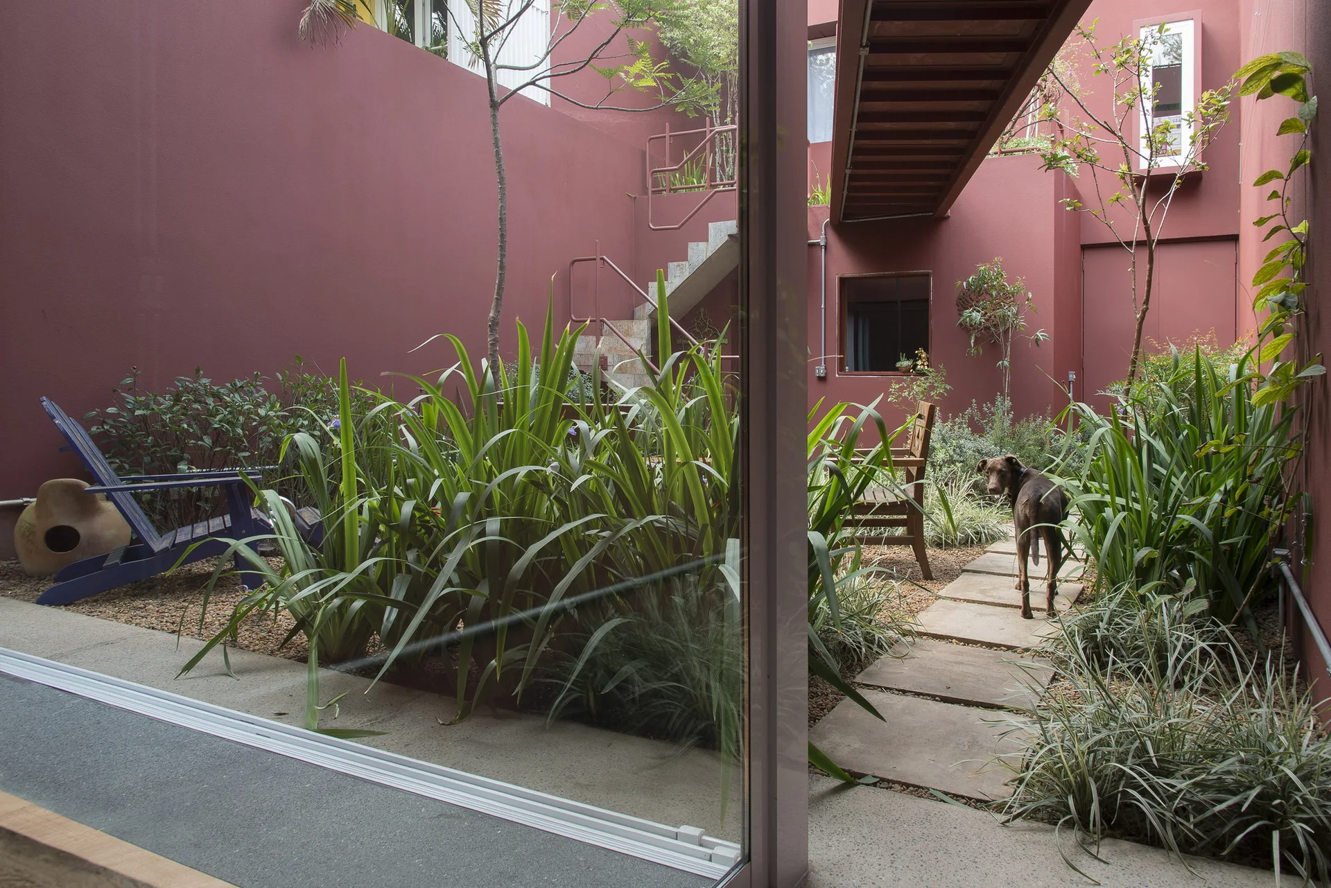 An enclosed modern courtyard features deep red walls, concrete stepping stones, lush green plants, and an overhead wooden walkway.