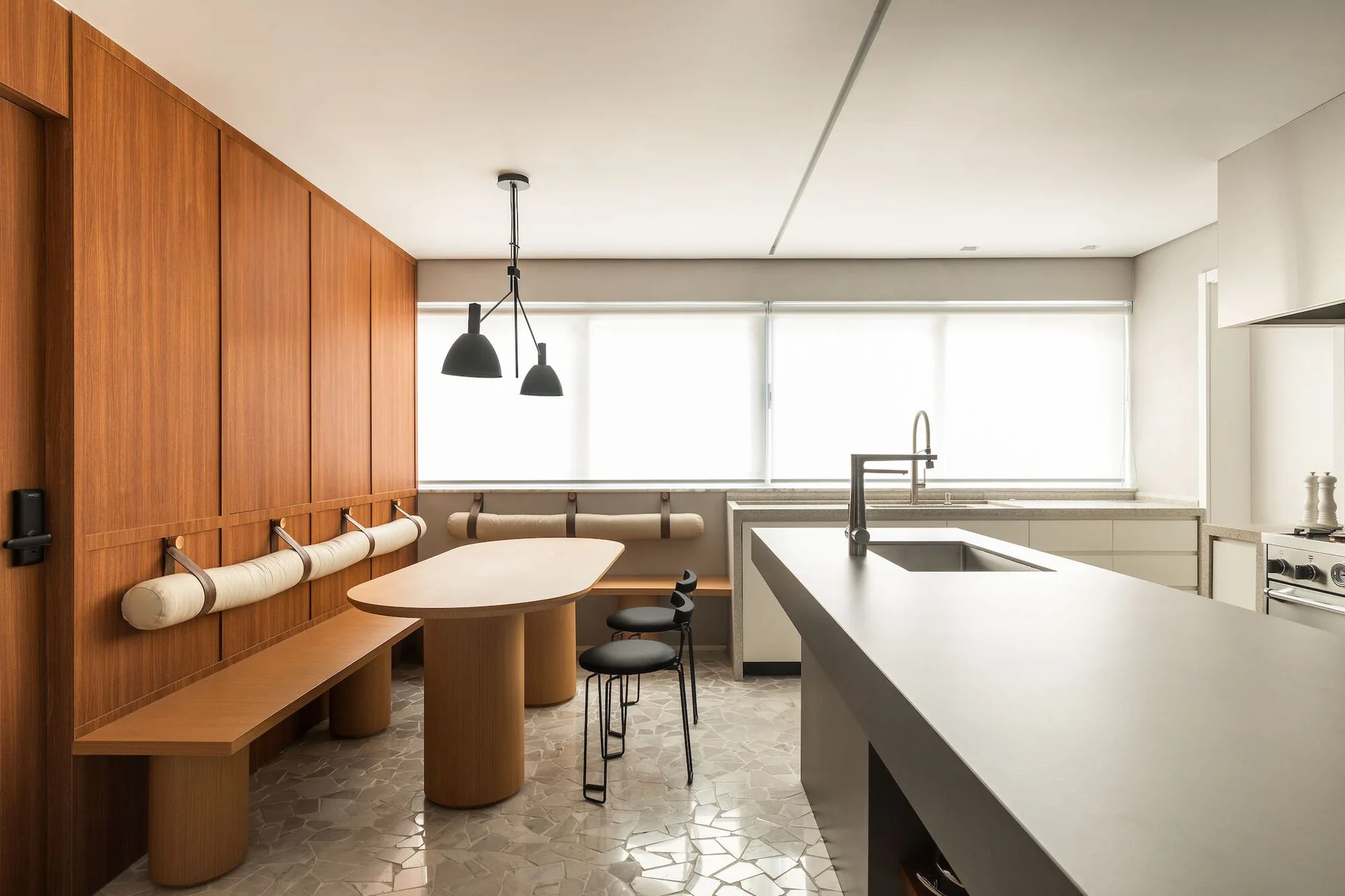 A modern kitchen featuring a grey stone island, an oval wood dining table with a built-in bench, and wall-mounted bolster cushions.