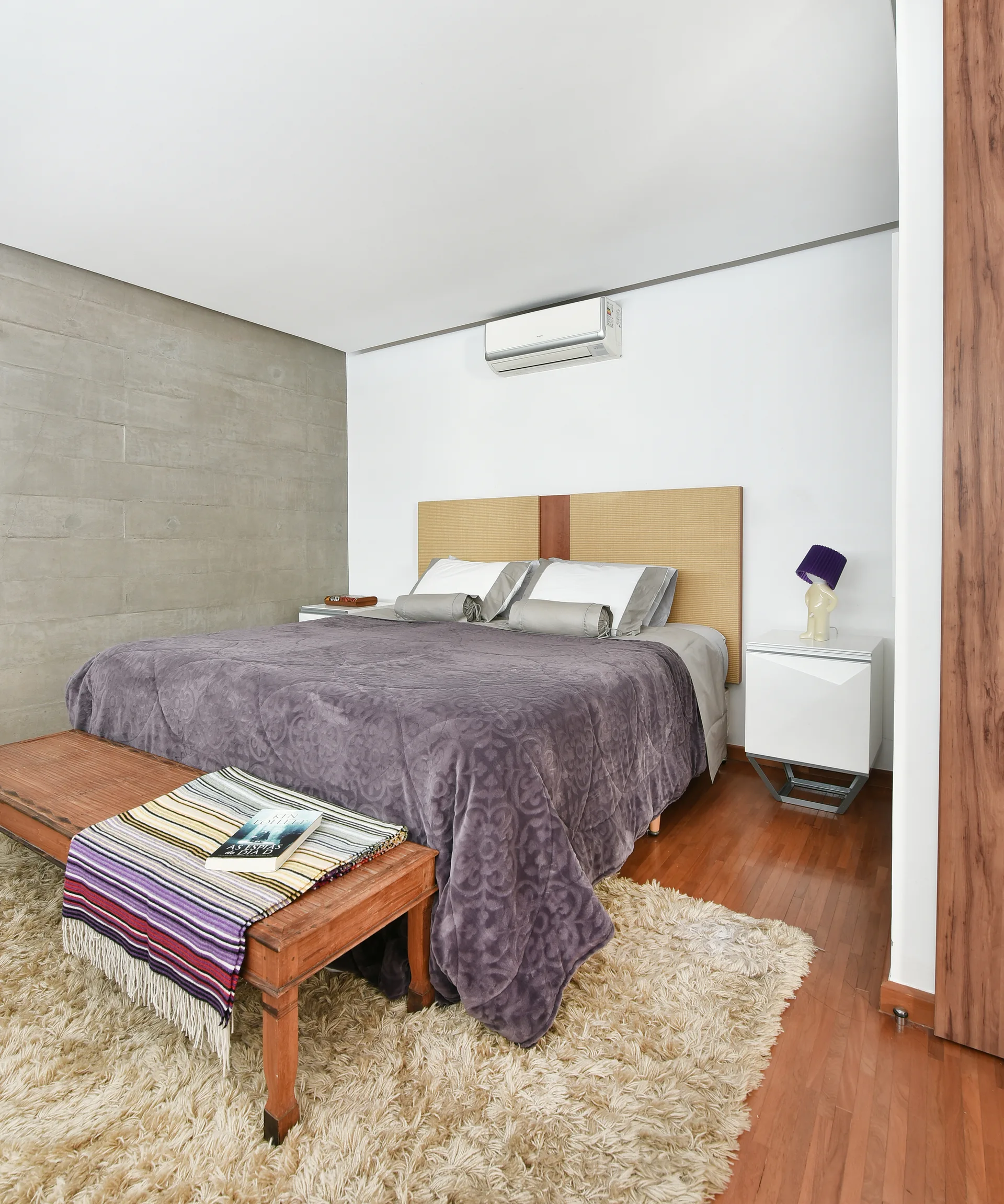 A modern bedroom featuring a bed with purple linens, an exposed concrete wall, a wooden bench, and a textured shag rug.