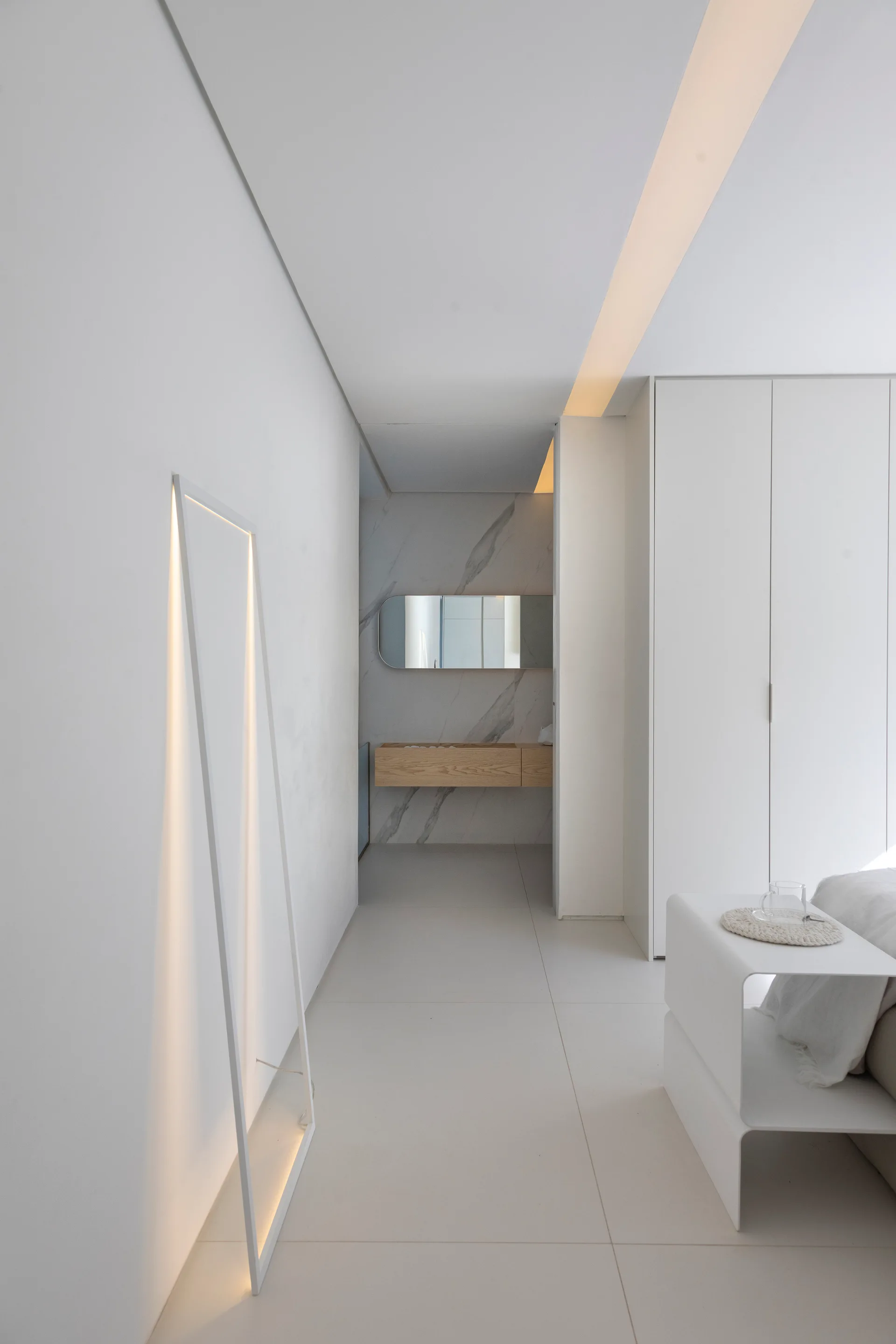 A white minimalist bedroom hallway features a leaning rectangular floor lamp, a built-in wardrobe, and a distant wooden floating vanity.
