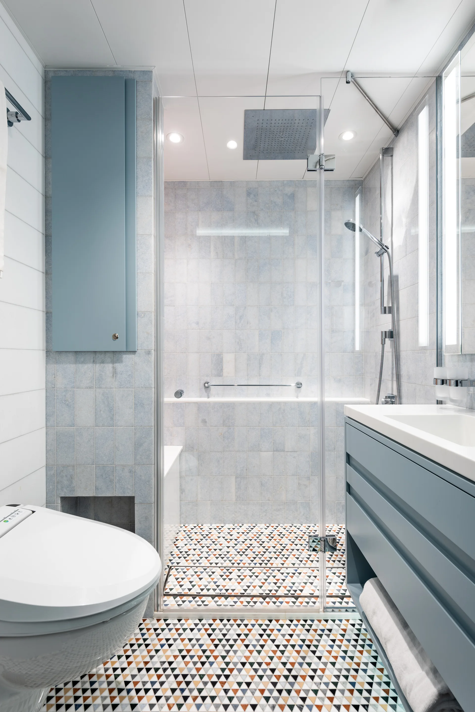 A modern bathroom featuring a blue floating vanity, a glass shower enclosure, and multicolored geometric floor tiles.
