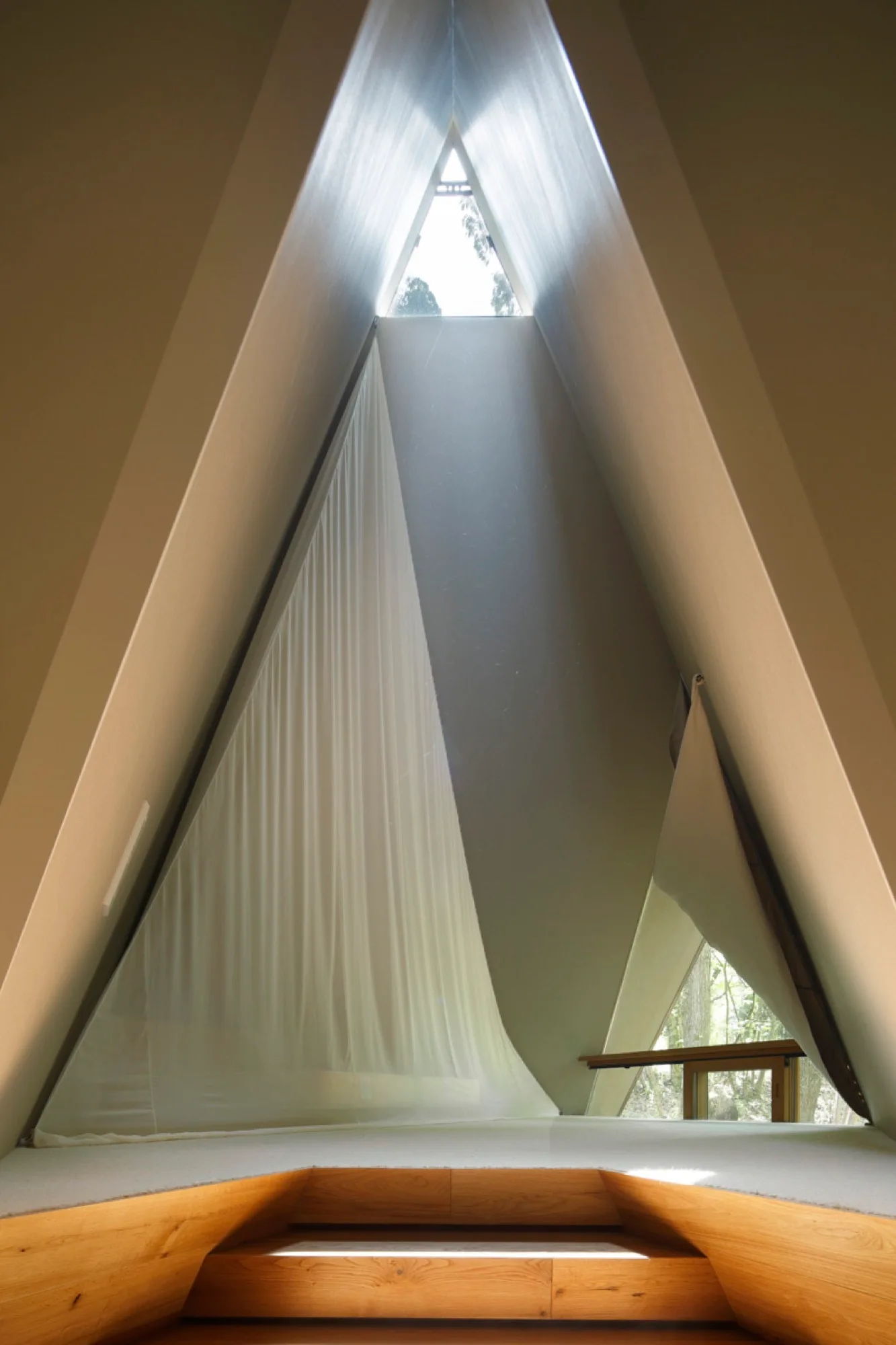 A minimalist A-frame interior featuring a high triangular skylight, off-white walls, a sheer white fabric canopy, and a stepped wooden platform.