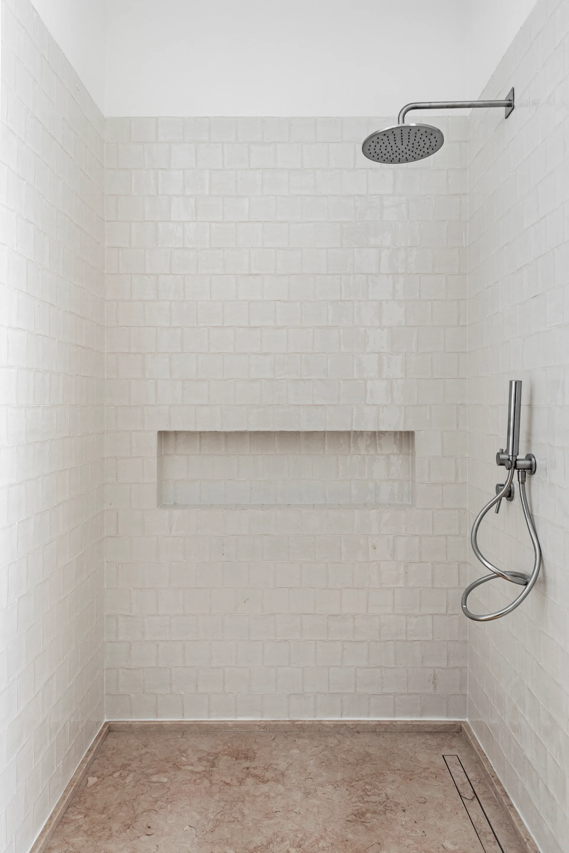 A modern walk-in shower featuring off-white square-tiled walls with a long recessed niche and a beige natural stone tray floor.