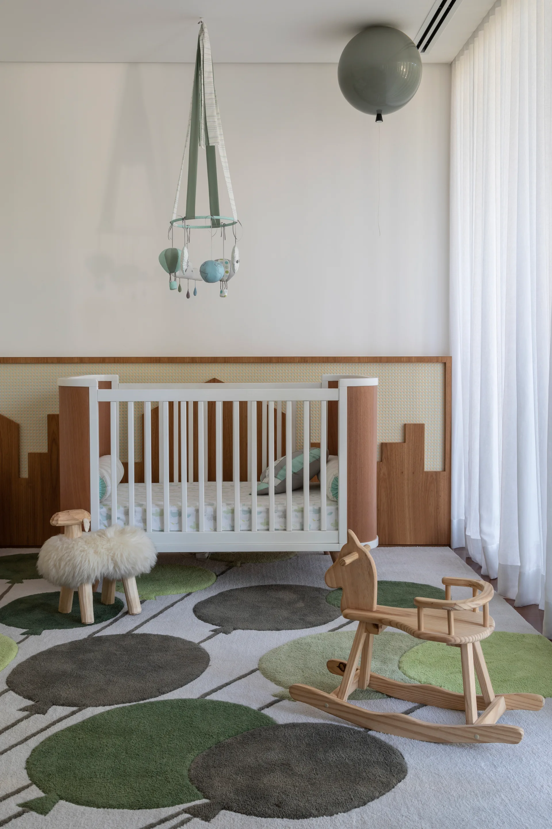 A modern nursery features a white and wood crib against cane wall paneling, with a balloon-patterned rug, rocking horse, and hanging mobile.
