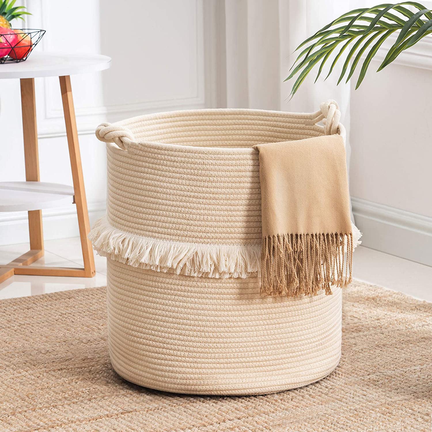 A cream woven rope basket with fringe details sits on a textured tan rug next to a white side table.