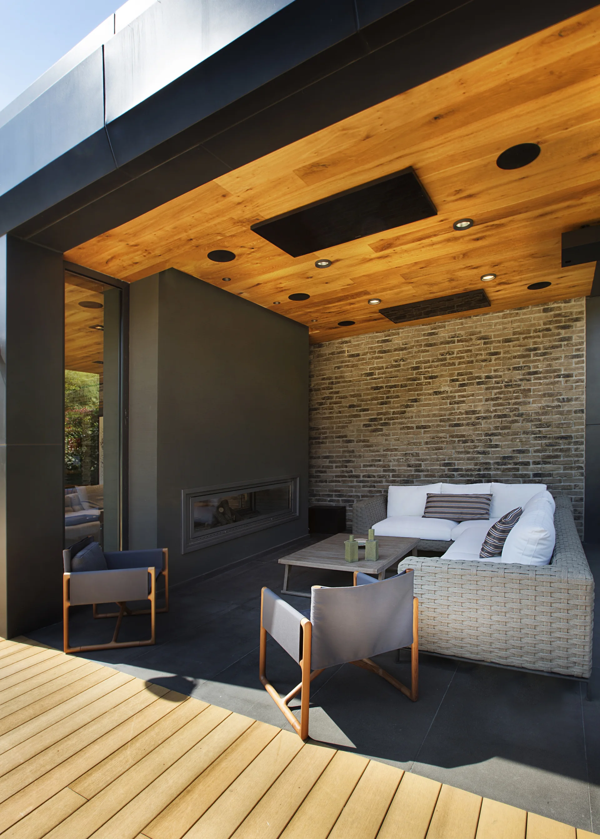 A contemporary covered patio featuring a wicker sectional sofa, two gray armchairs, a built-in linear fireplace, and a warm wood-paneled ceiling.