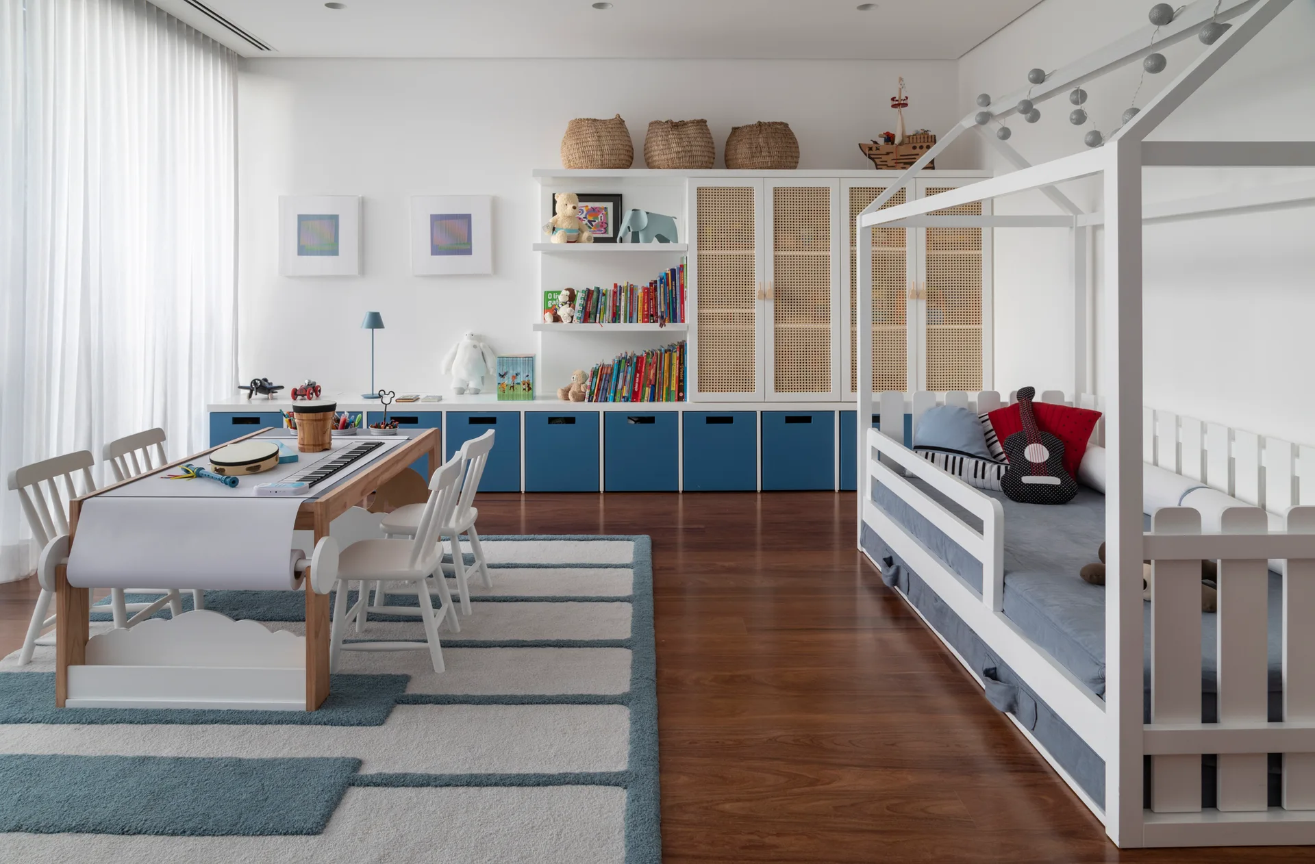 A children's playroom with a white house-shaped bed frame, blue and white built-in storage cabinets, and a long art desk on a striped rug.
