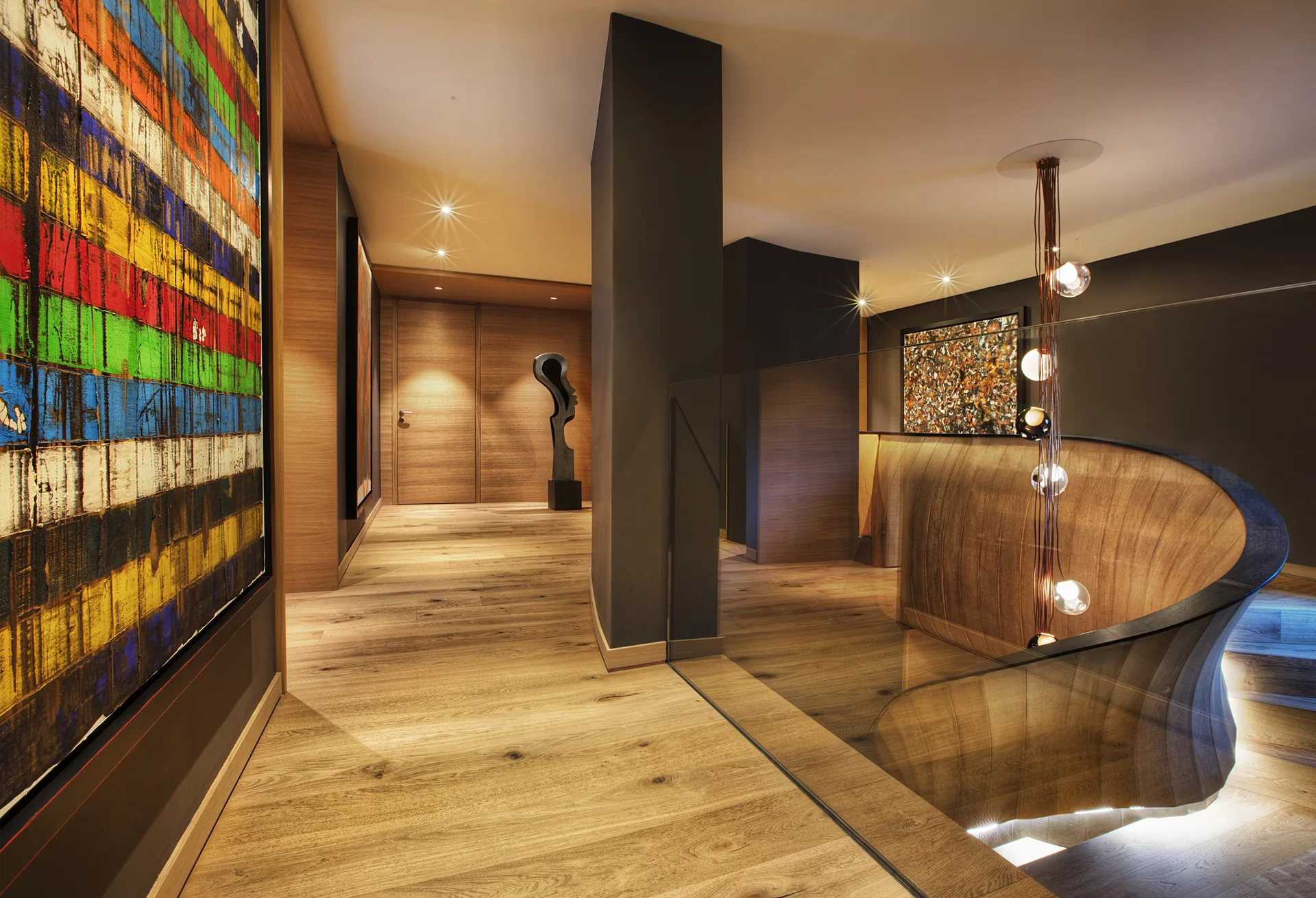 A contemporary hallway landing features a curved wood staircase with a glass railing, dark charcoal accent walls, and wide-plank wood flooring.