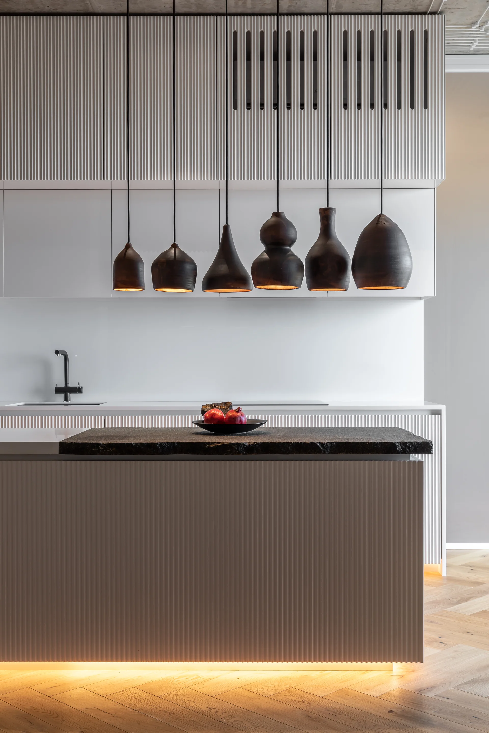 A minimalist kitchen features a white fluted island with a raw-edge dark stone countertop, lit by six black organic-shaped pendant lights.