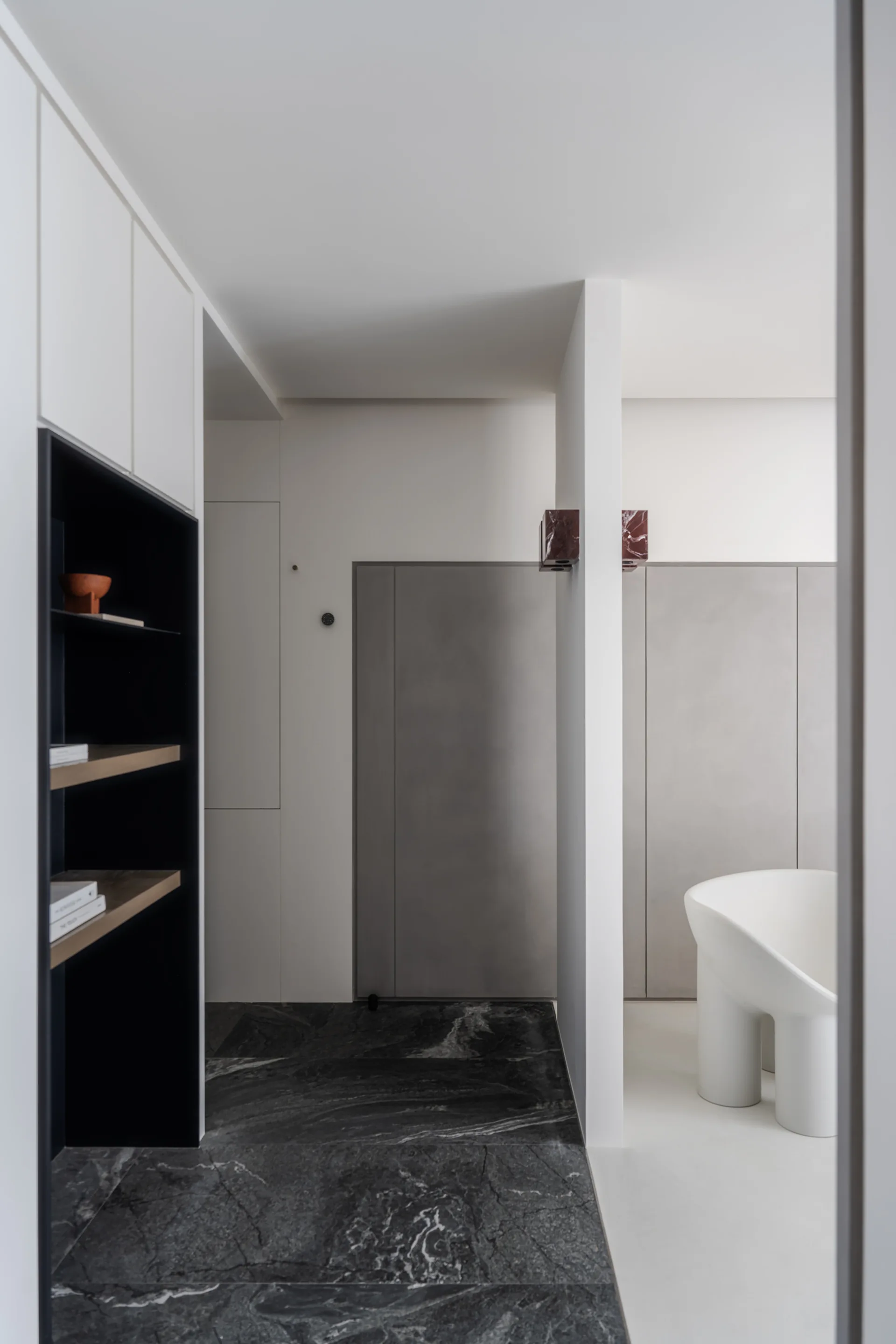 A minimalist hallway featuring dark veined marble flooring, flush white and grey cabinetry, built-in black shelving, and a sculptural white chair.