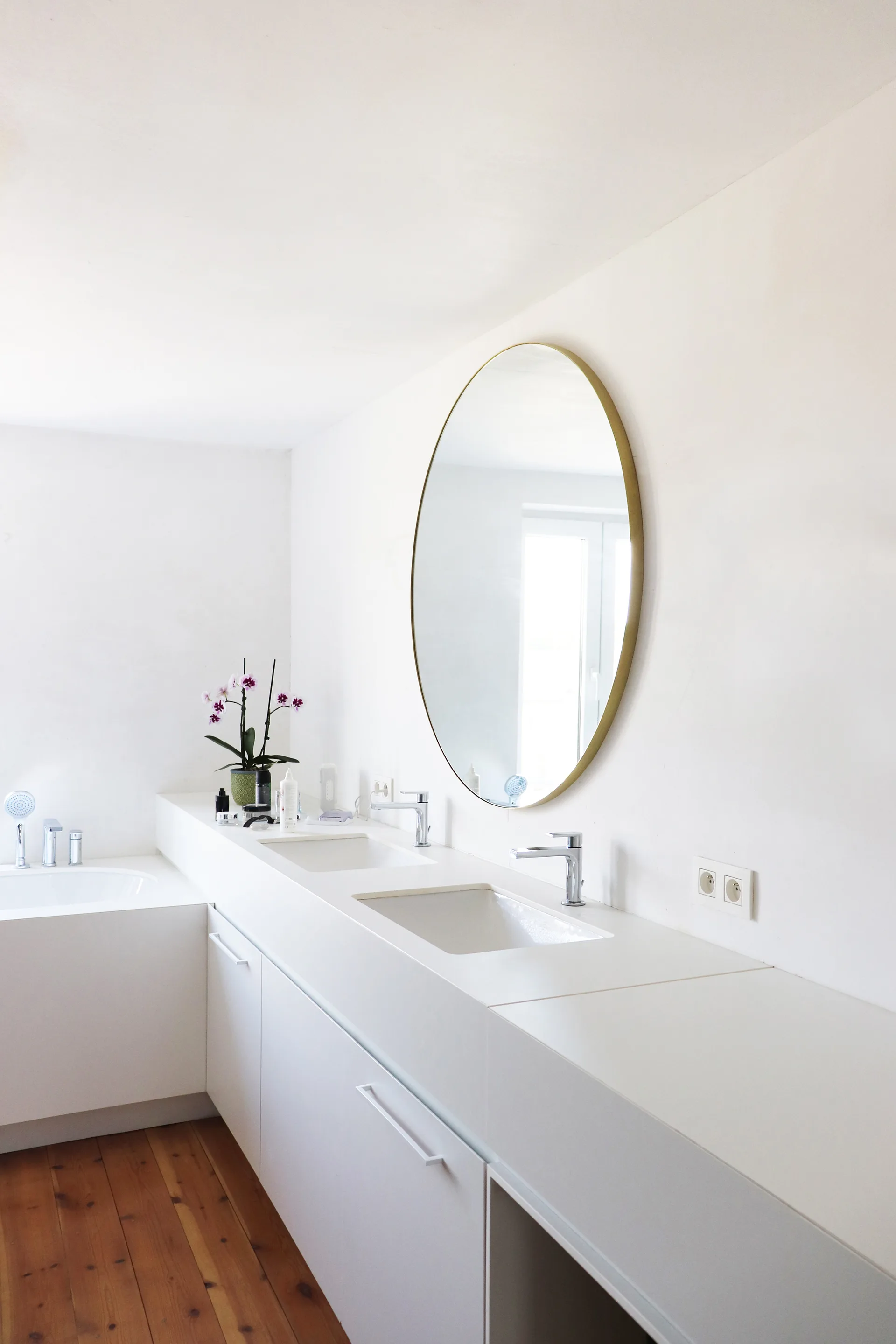 A minimalist white bathroom featuring a long double-sink vanity, a large round mirror, a built-in bathtub, and warm wooden plank flooring.