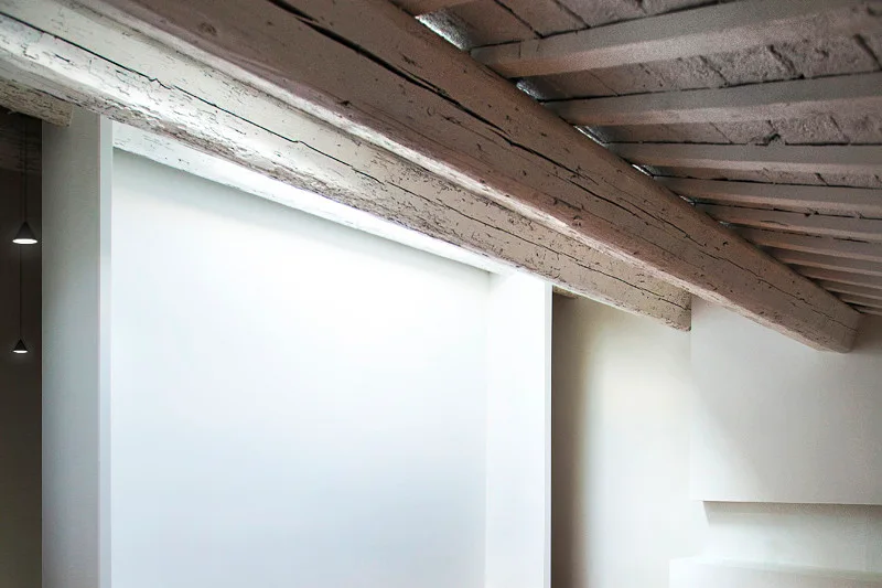 A rustic weathered wood ceiling beam contrasts against a smooth white wall illuminated by indirect recessed architectural lighting.