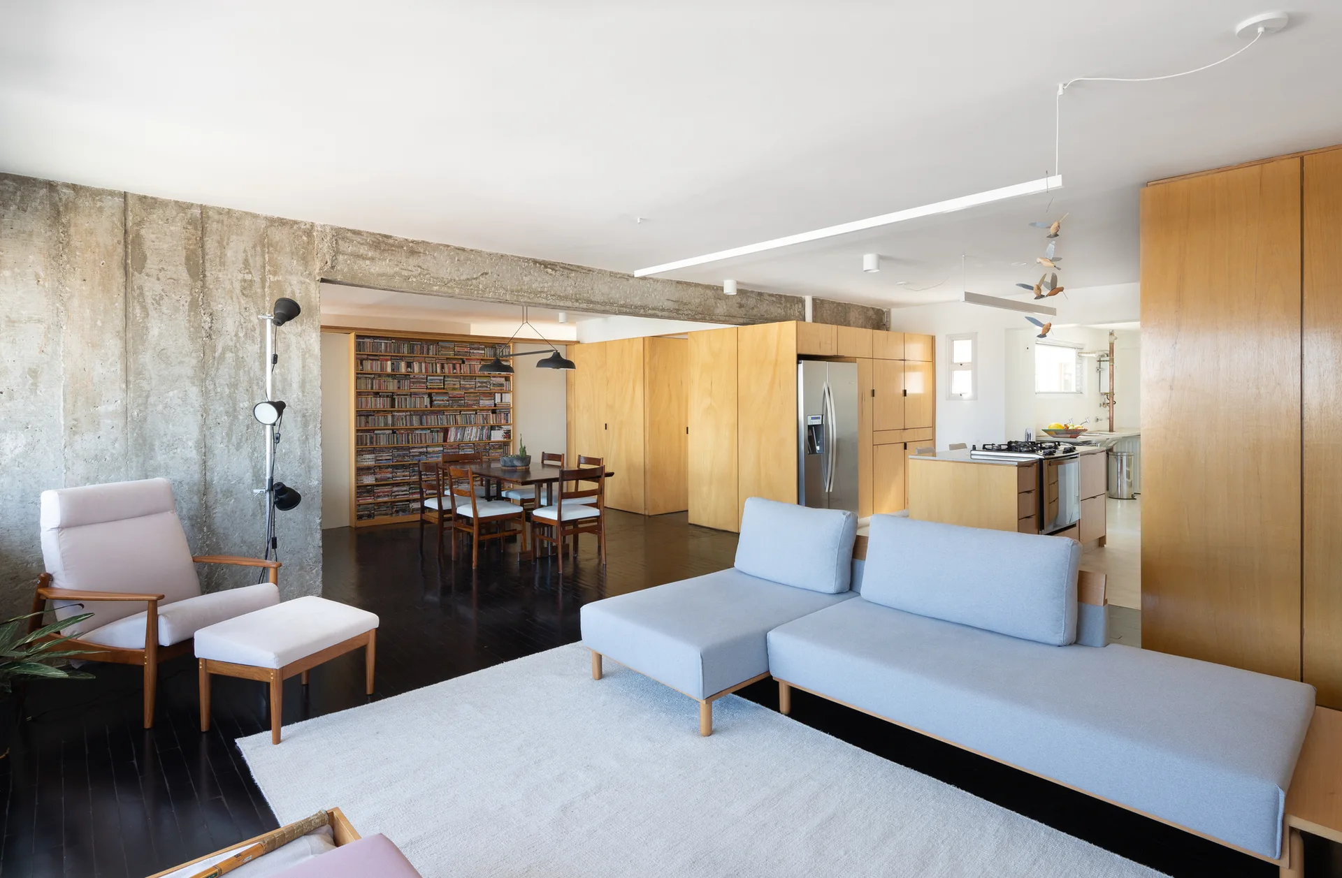 An open-plan living room features a grey sectional sofa, a plywood wall system, an exposed concrete beam, and a dining area in the background.