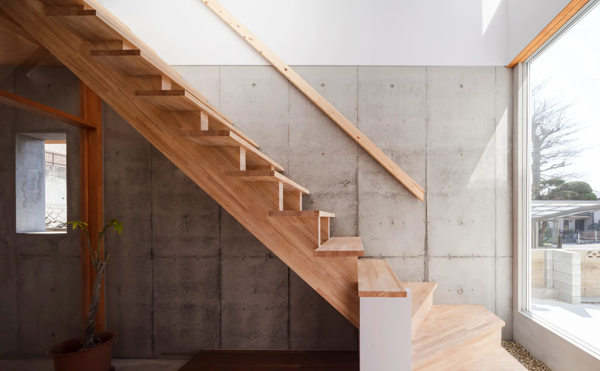 An open-riser wooden staircase ascends alongside a textured concrete wall next to a large window bringing in natural light.