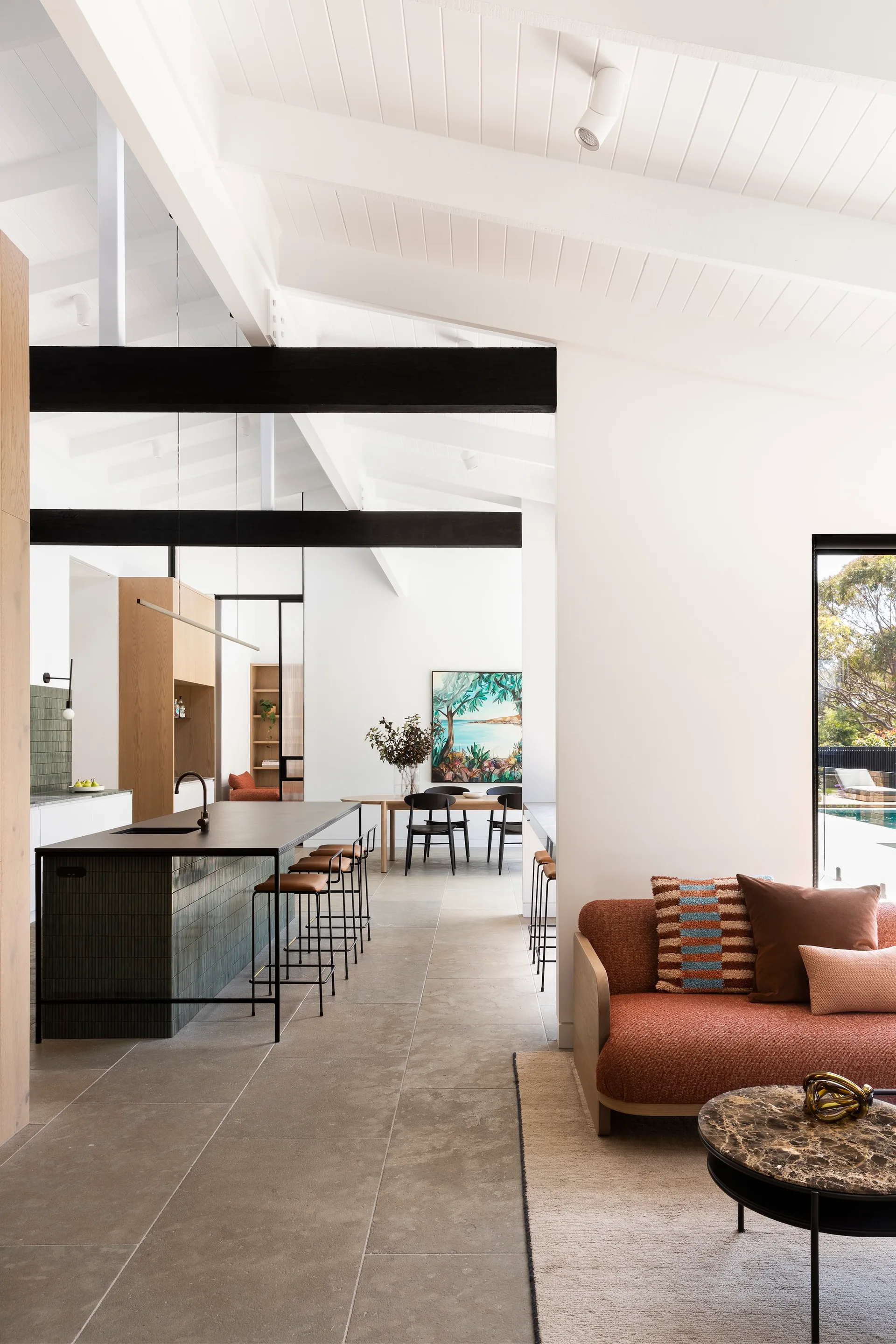An open-plan living space features a terracotta sofa, a dark-topped kitchen island, and a vaulted ceiling with exposed black beams.