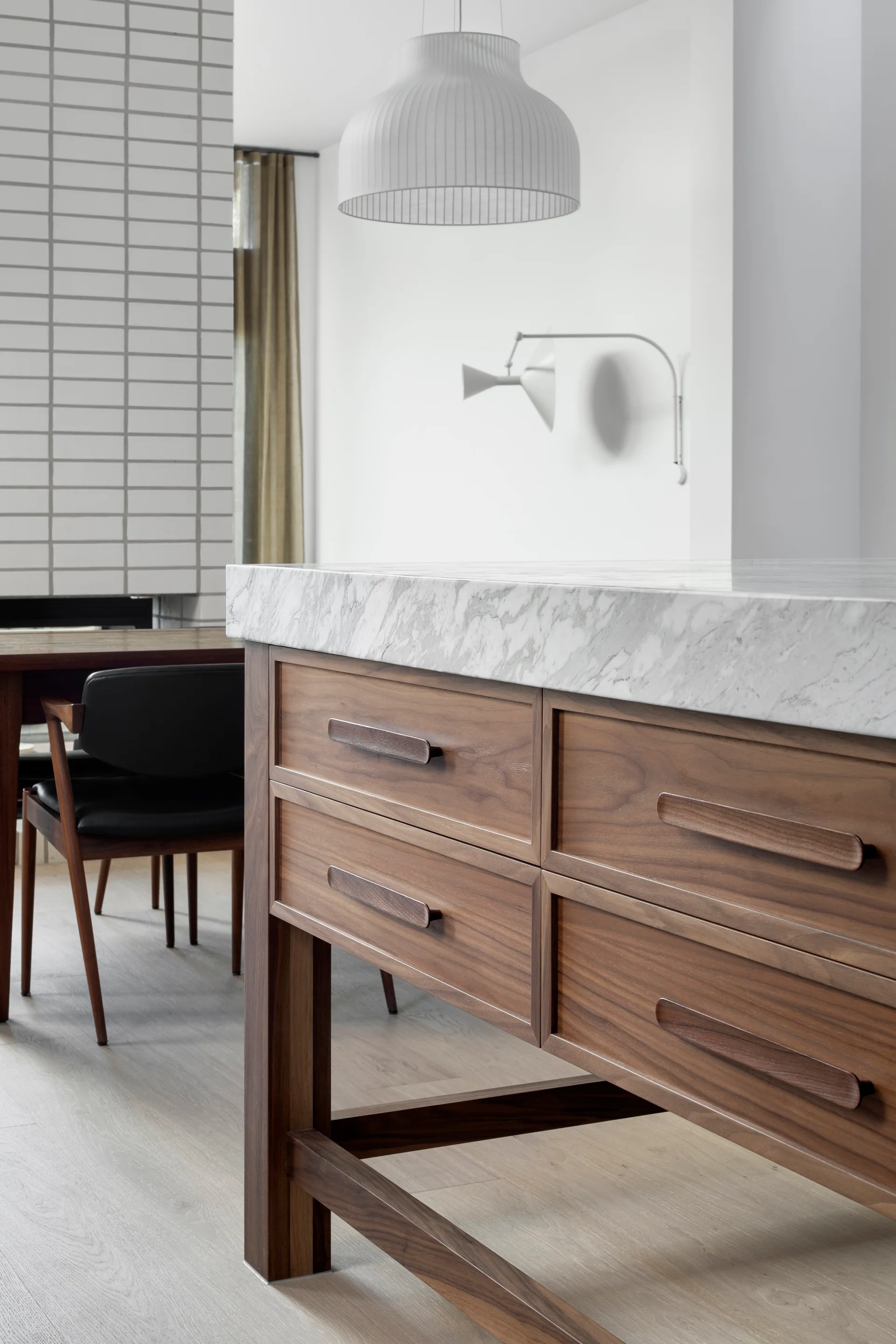 A close-up of a modern walnut kitchen island featuring a marble countertop and integrated wood drawer handles, with a dining room in the background.