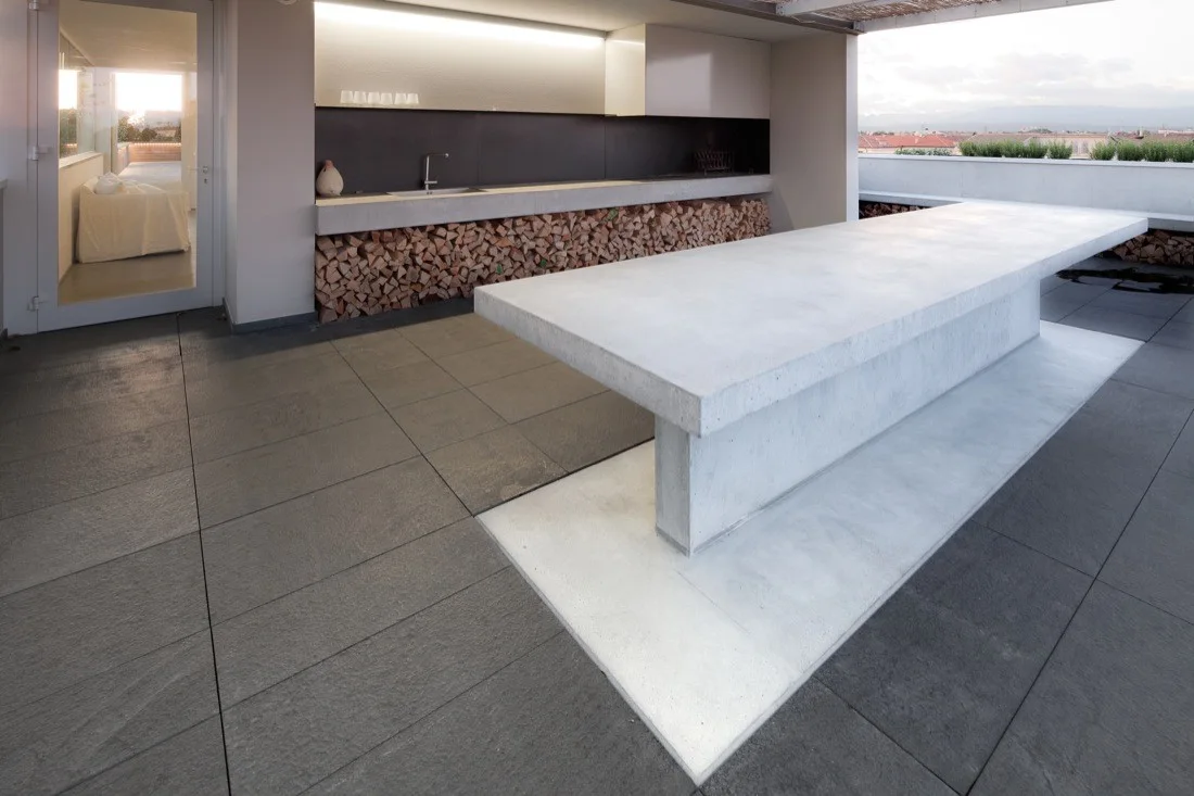 A minimalist outdoor kitchen on a rooftop terrace features a large concrete dining island and a long prep counter with stacked logs underneath.