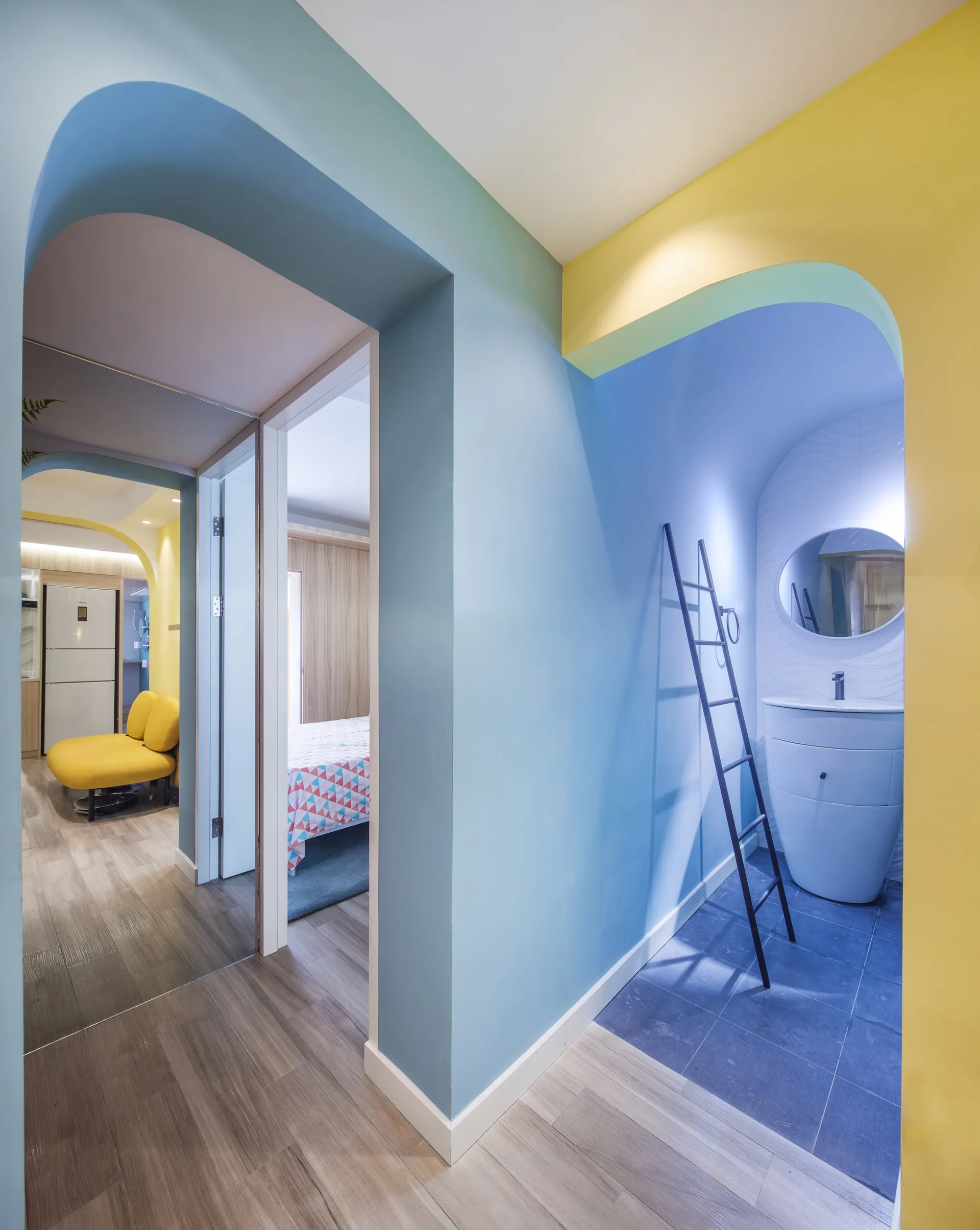 A residential hallway features arched doorways painted blue and yellow leading to a bathroom with a white vanity and a bedroom with wood floors.