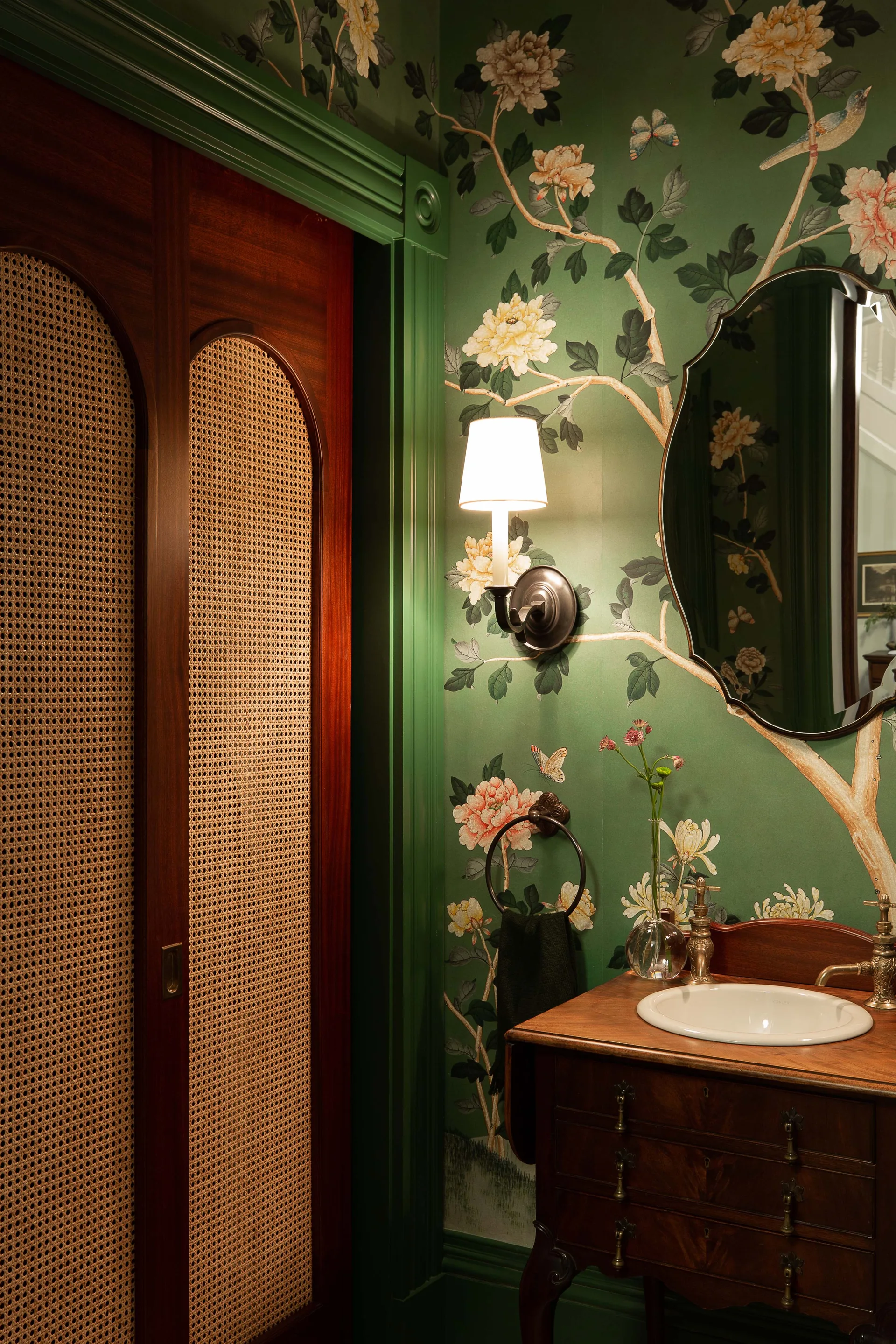 A powder room features green botanical wallpaper, a dark wood vanity with a brass faucet, a shaped mirror, and a wood sliding door with woven cane inserts.