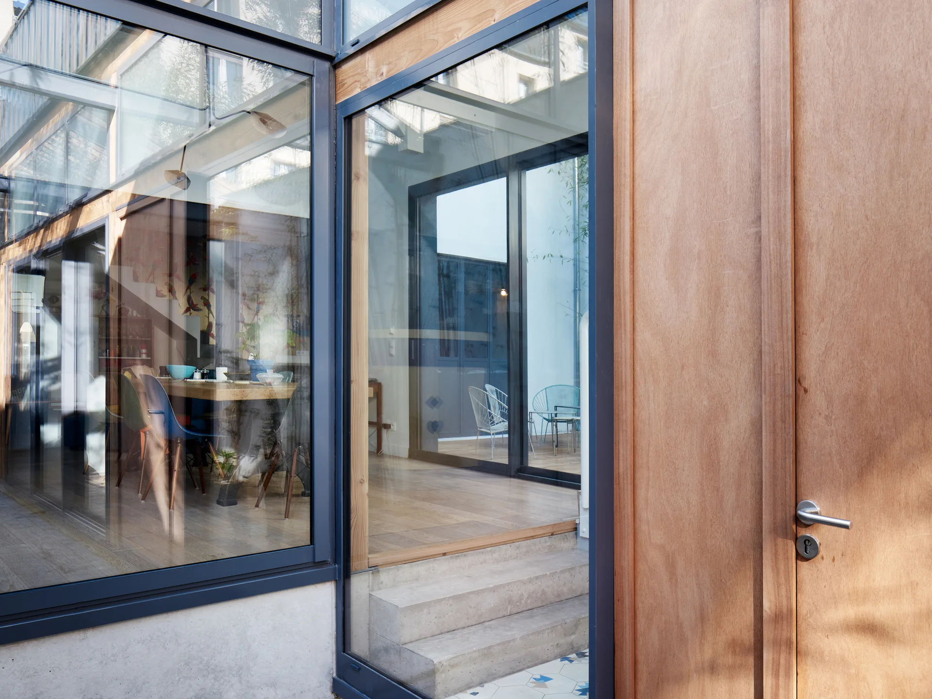 A modern home entrance features large glass walls, a light wood door frame, concrete steps, and a glimpse of an indoor dining room.