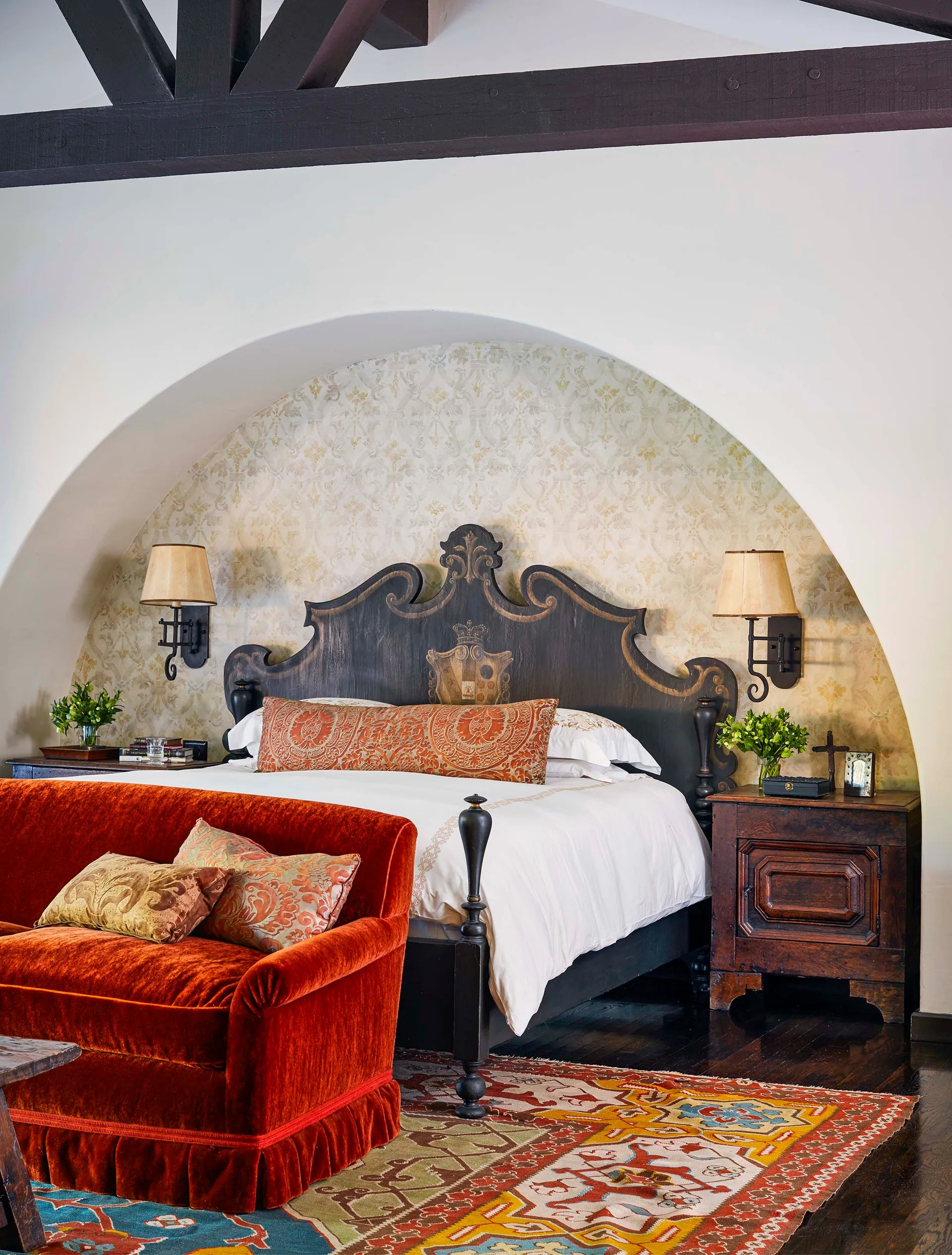 A traditional bedroom features an ornate dark wood bed framed by an arched wallpapered alcove, alongside a bright orange velvet sofa.