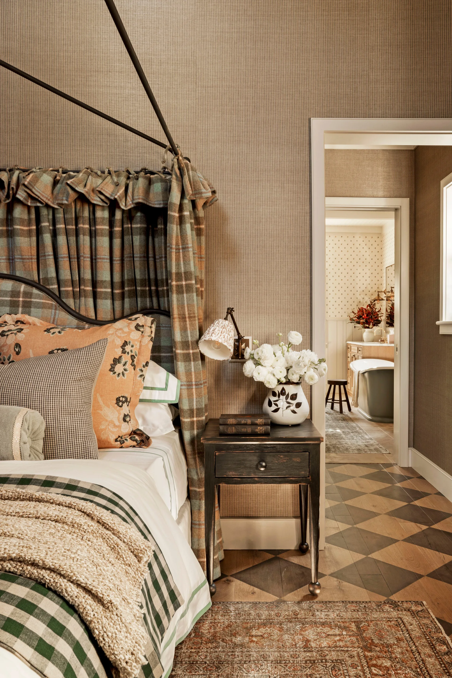 A bedroom features a black metal four-poster bed with plaid drapery next to a distressed dark nightstand, with a view into an adjacent bathroom.