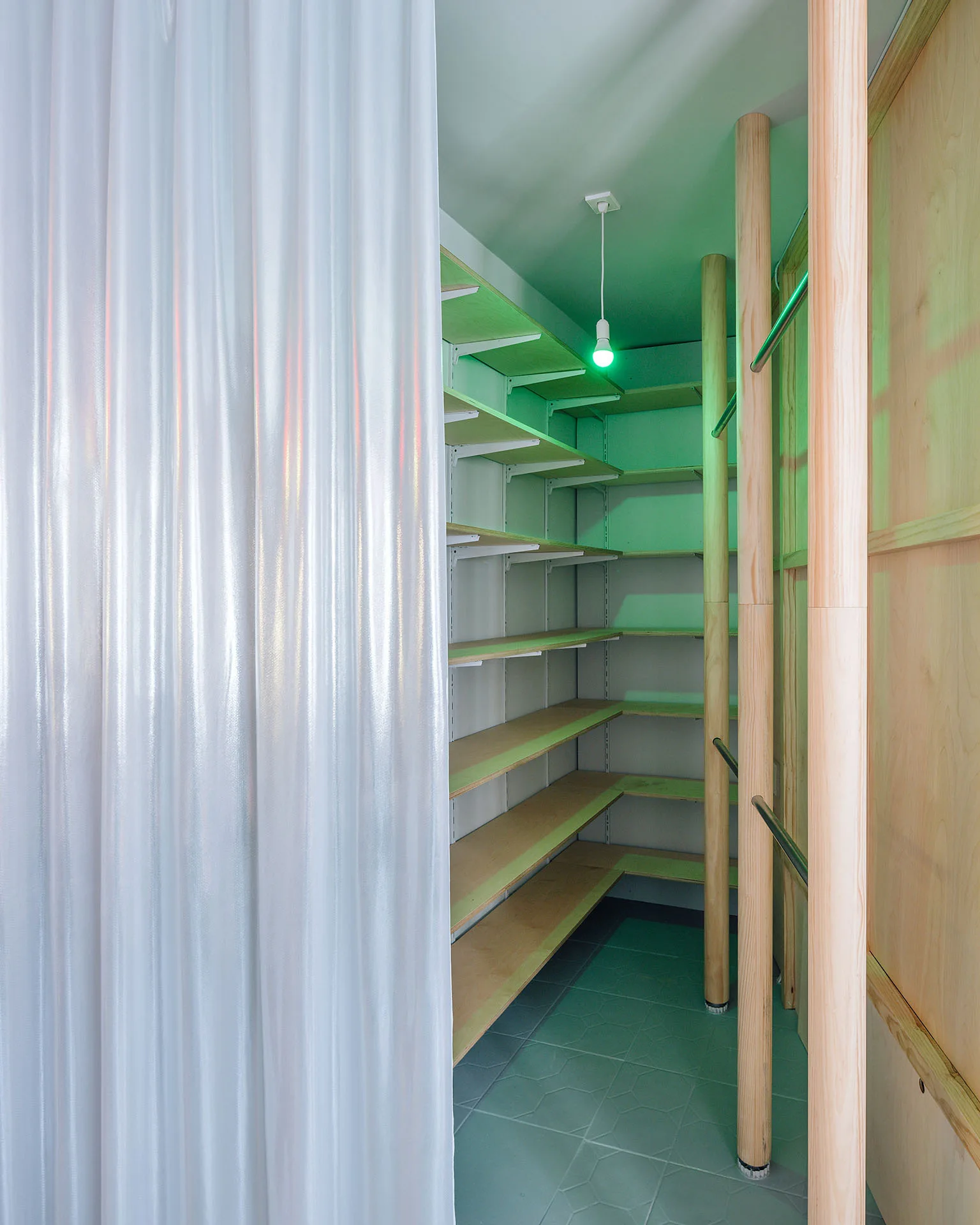 An interior storage closet features open wood shelving, vertical wood pillars, and a translucent white plastic curtain partition under a green light bulb.
