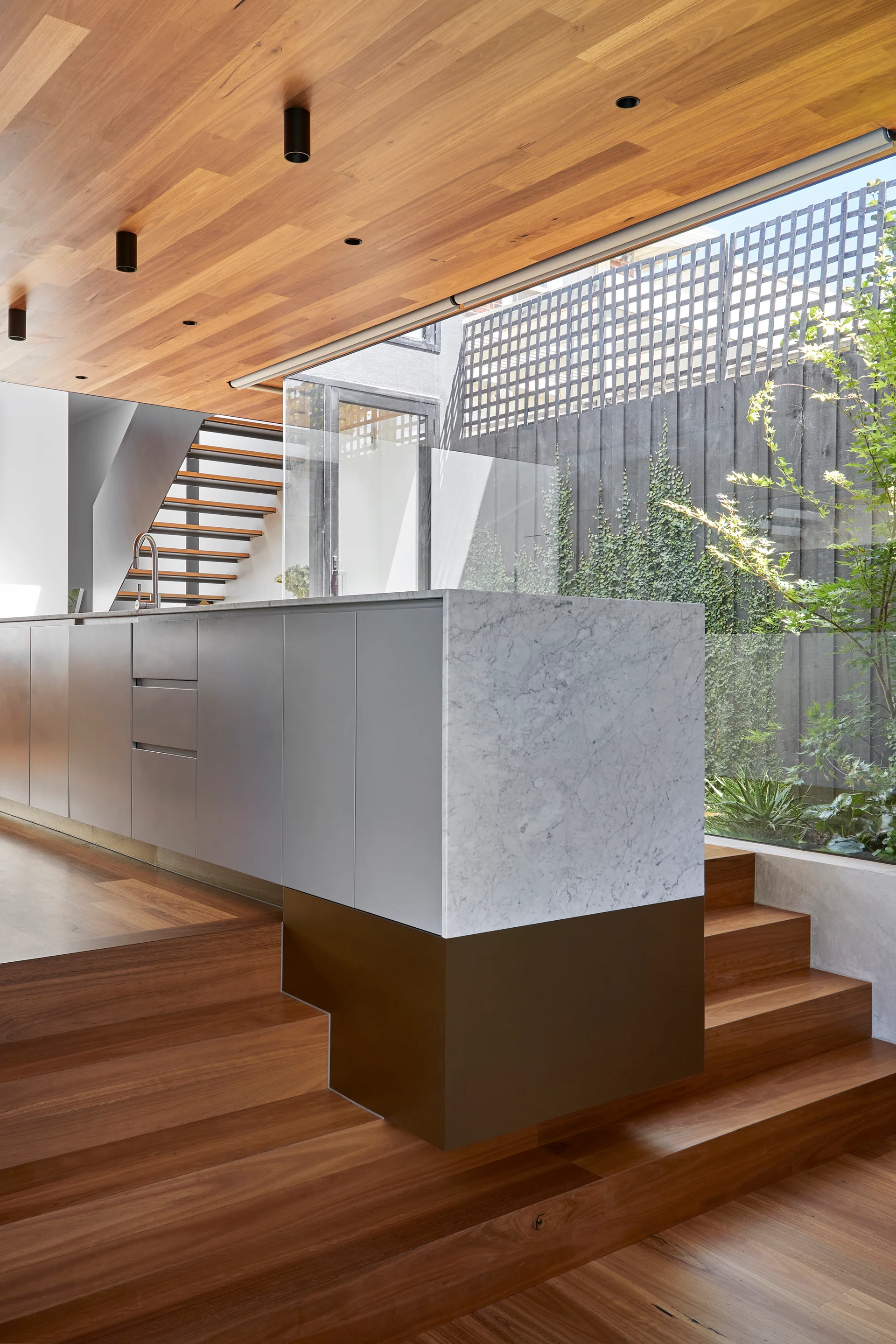 A minimalist kitchen features a marble-topped island on tiered wood stairs, alongside a wood ceiling and a window looking out onto a garden.