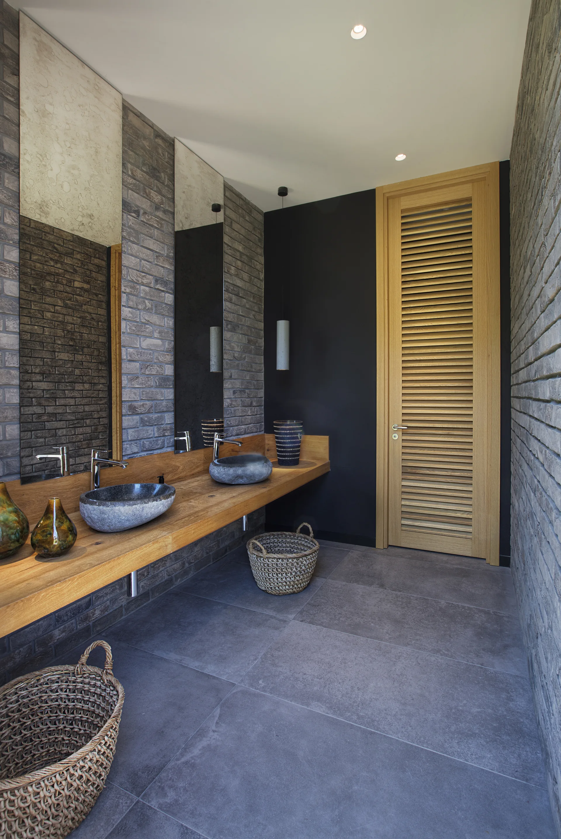 A contemporary bathroom featuring two grey stone vessel sinks on a wooden floating vanity next to a textured grey brick wall.