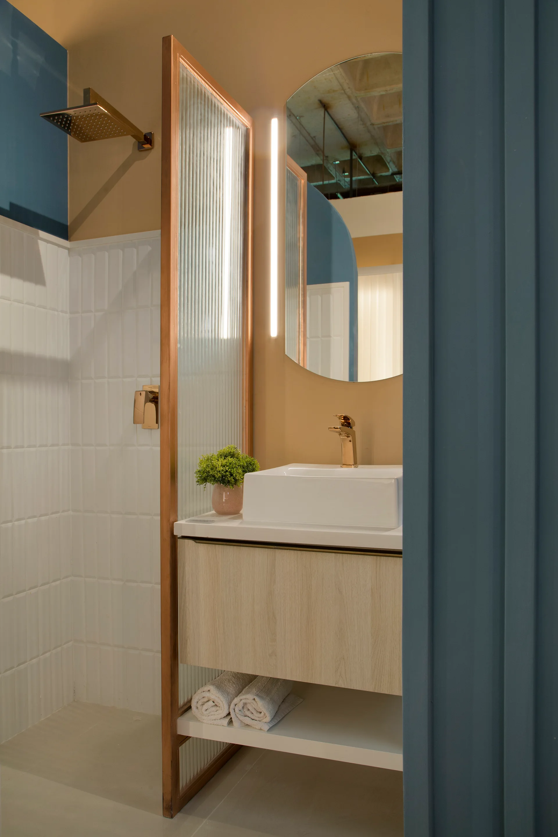 A contemporary bathroom with a light wood vanity, white vessel sink, gold fixtures, and a ribbed glass partition.