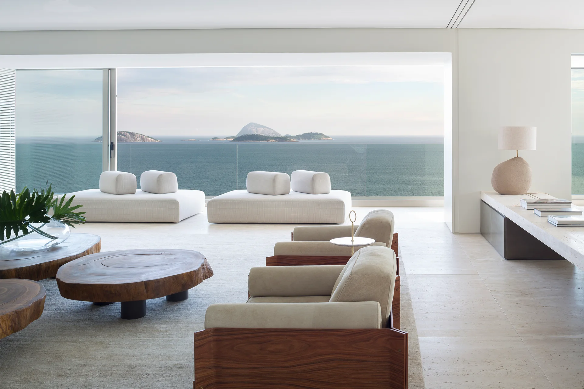 A minimalist living room with low white sofas, wooden armchairs, and live-edge coffee tables overlooking a vast ocean view.