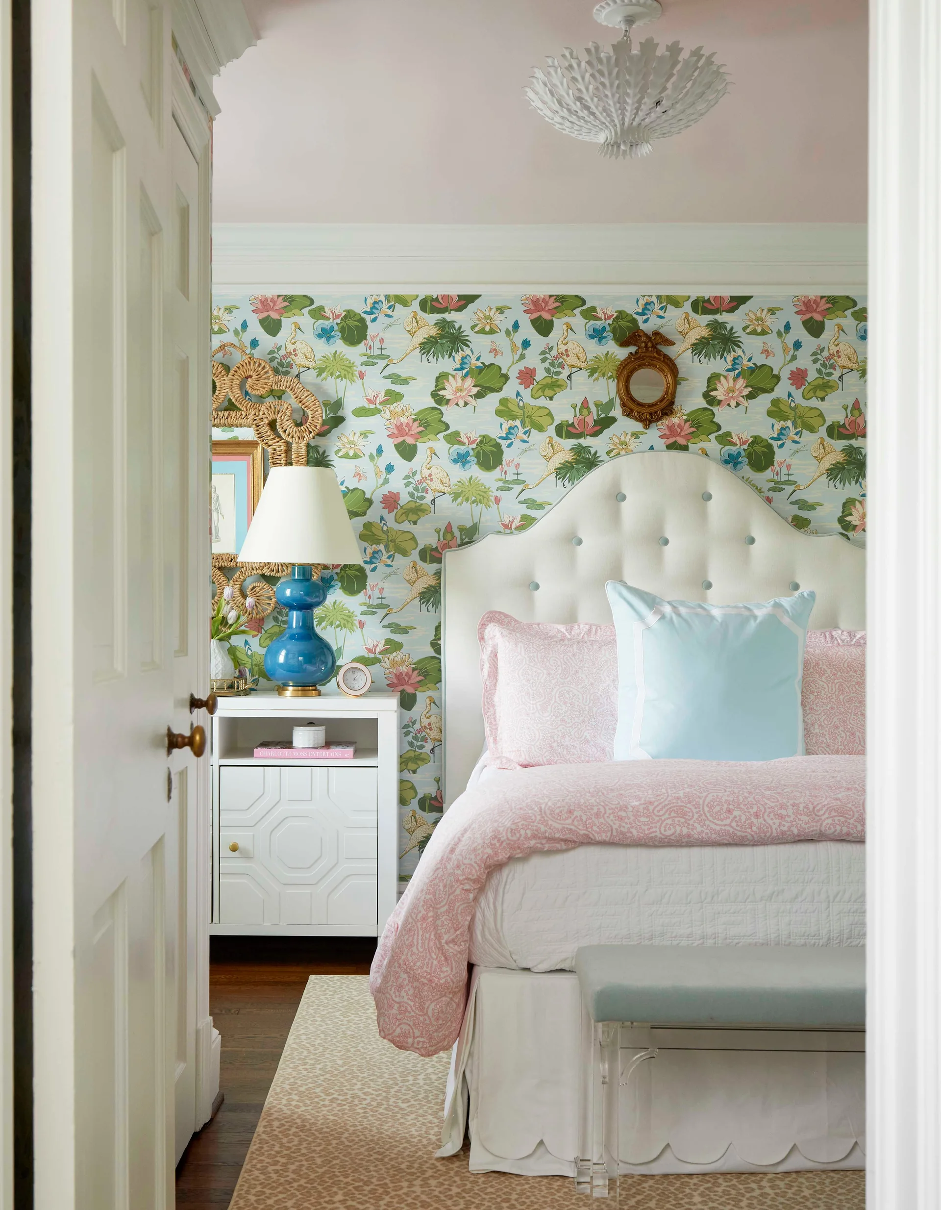 A bedroom viewed through a doorway features colorful botanical wallpaper, a white tufted headboard, pink bedding, and a blue glass lamp.