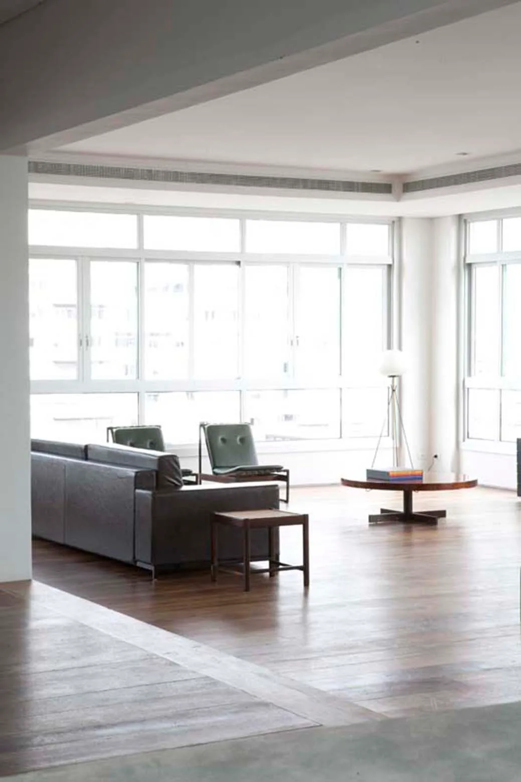 A minimalist living room features a dark leather sofa, two green armchairs, and a round wooden coffee table set against large windows.