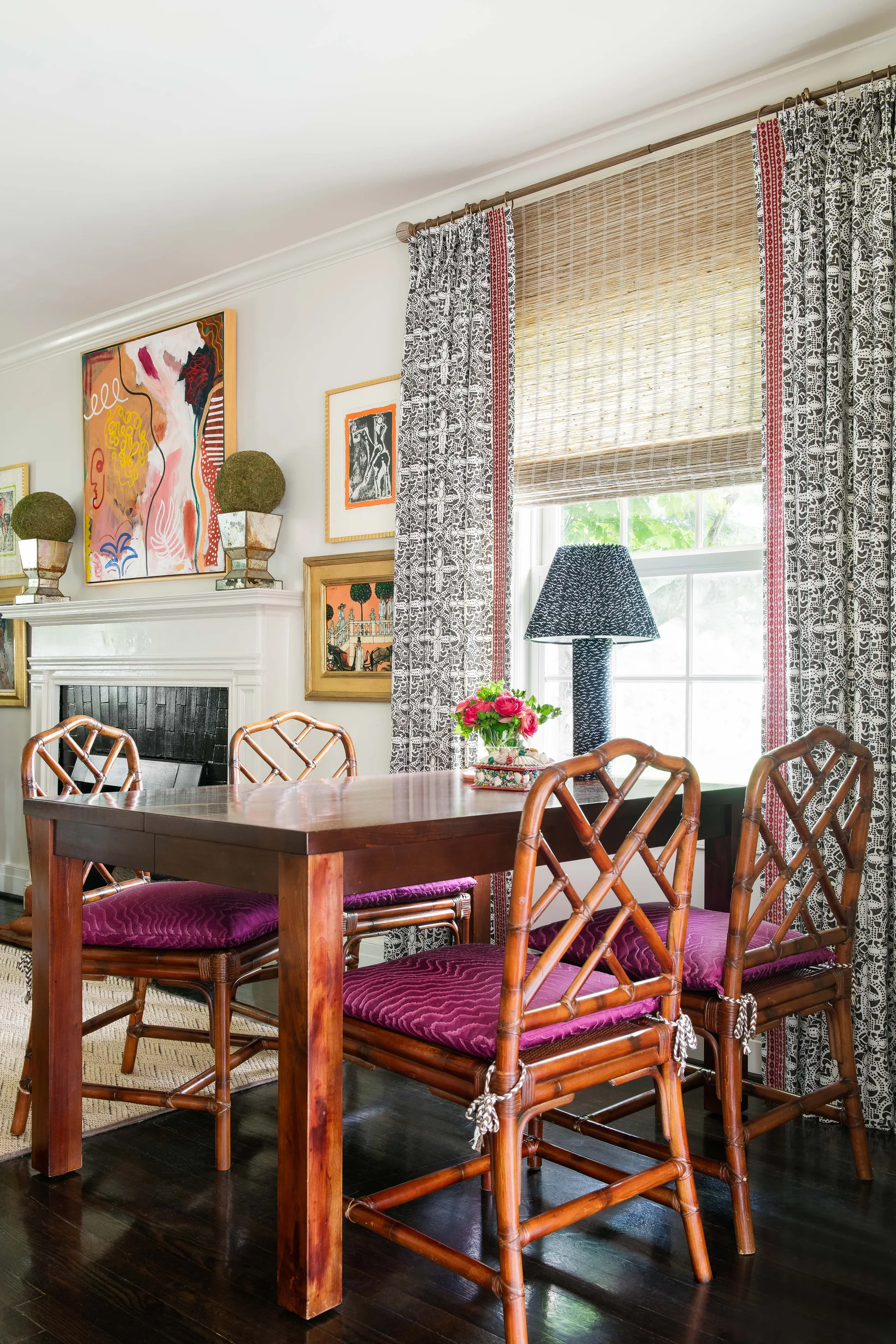 A dark wood dining table surrounded by bamboo chairs with magenta cushions sits in front of a fireplace and a window with patterned curtains.